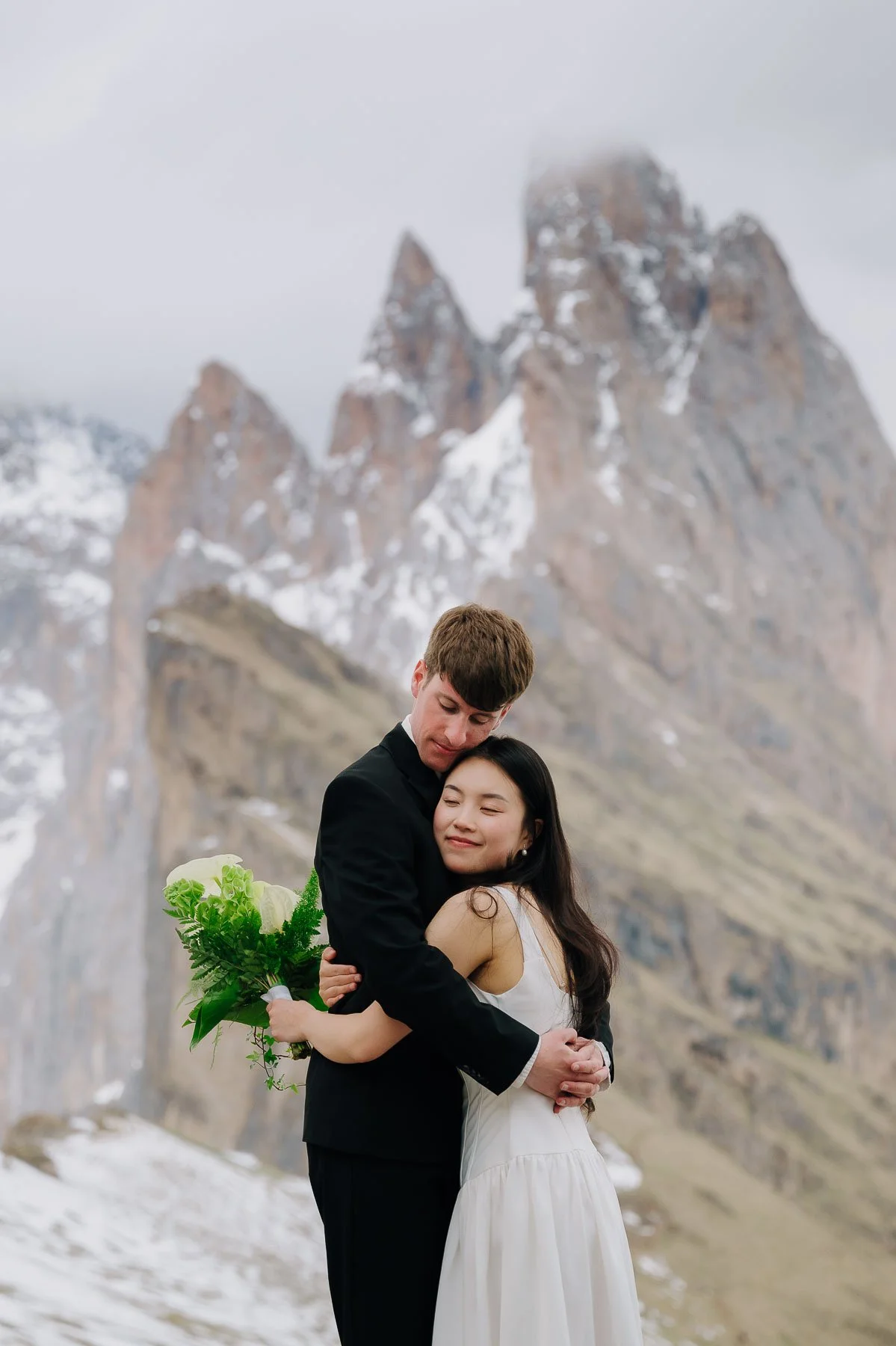A couple embracing outdoors with snowy mountains in the background, the woman holding a bouquet of flowers.