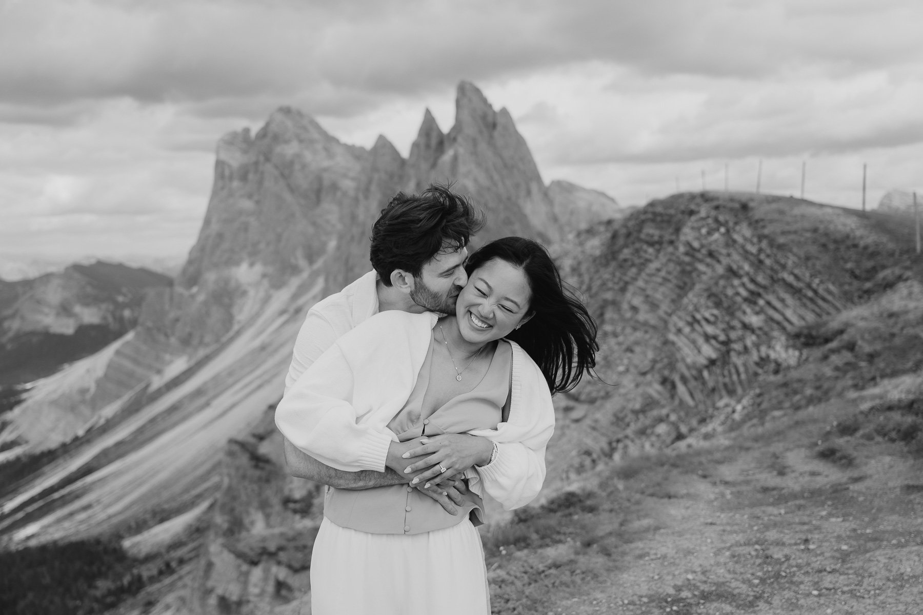 A black-and-white photo of a happy couple embracing in an outdoor mountainous landscape with rocky peaks in the background. The man is kissing the woman on the cheek while she smiles.