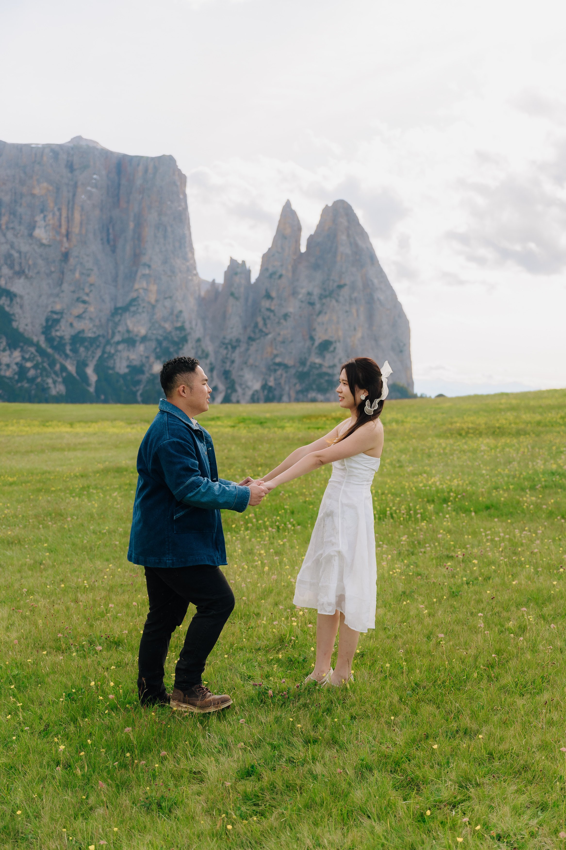 A man and woman standing in a green field holding hands, with mountains in the background, gazing at each other.