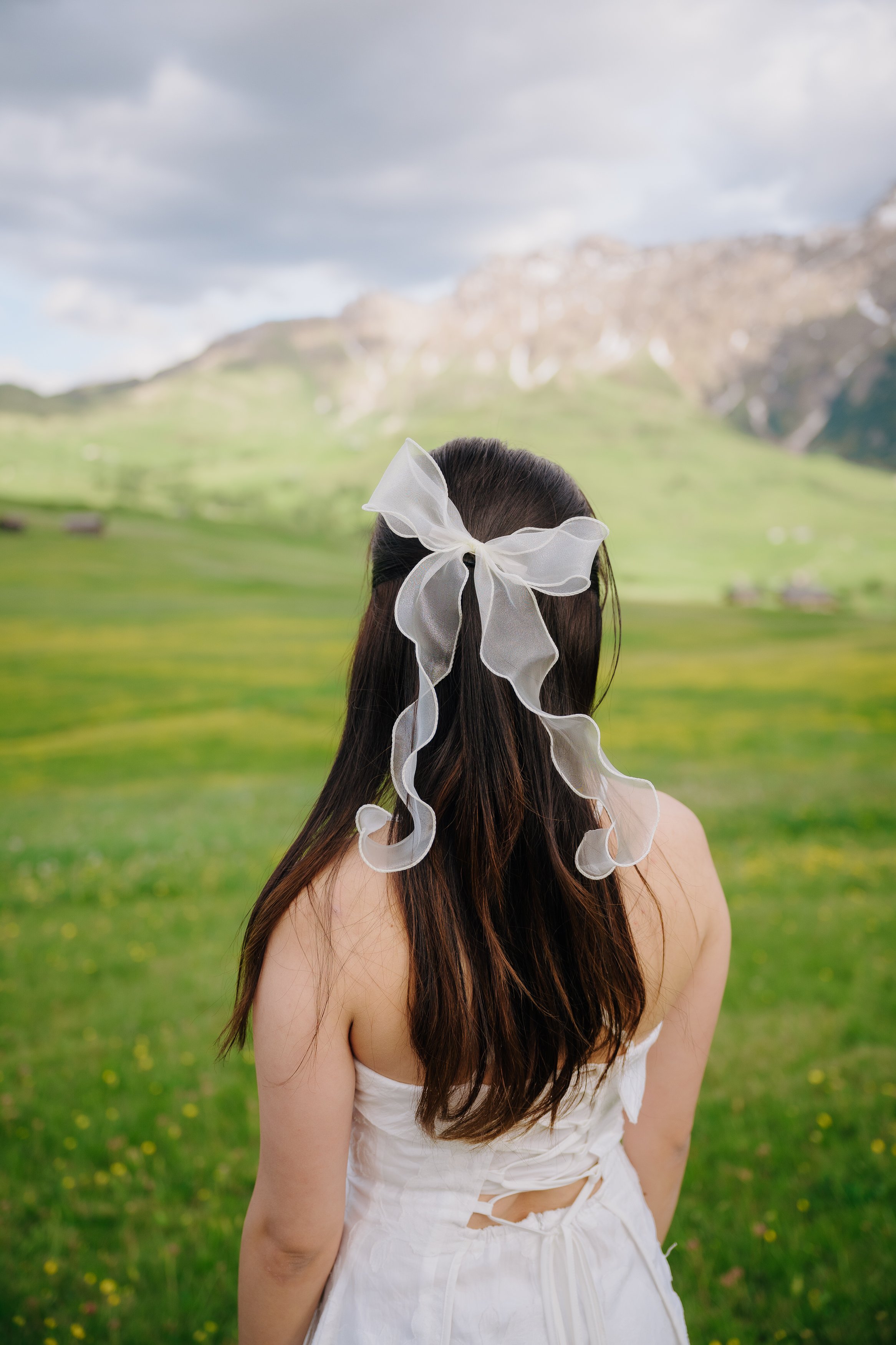 A woman with long dark hair, viewed from behind, wearing a white ribbon in her hair and a white dress, standing outdoors in a grassy field with mountains and cloudy sky in the background.