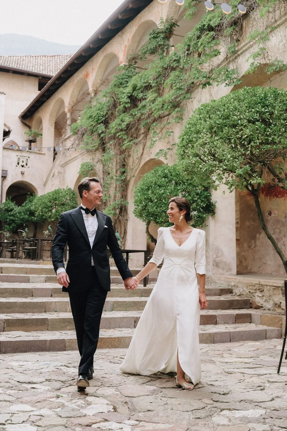 A couple in wedding attire walking hand in hand in a courtyard with stone steps, lush green trees, and a historic building with arches in the background.