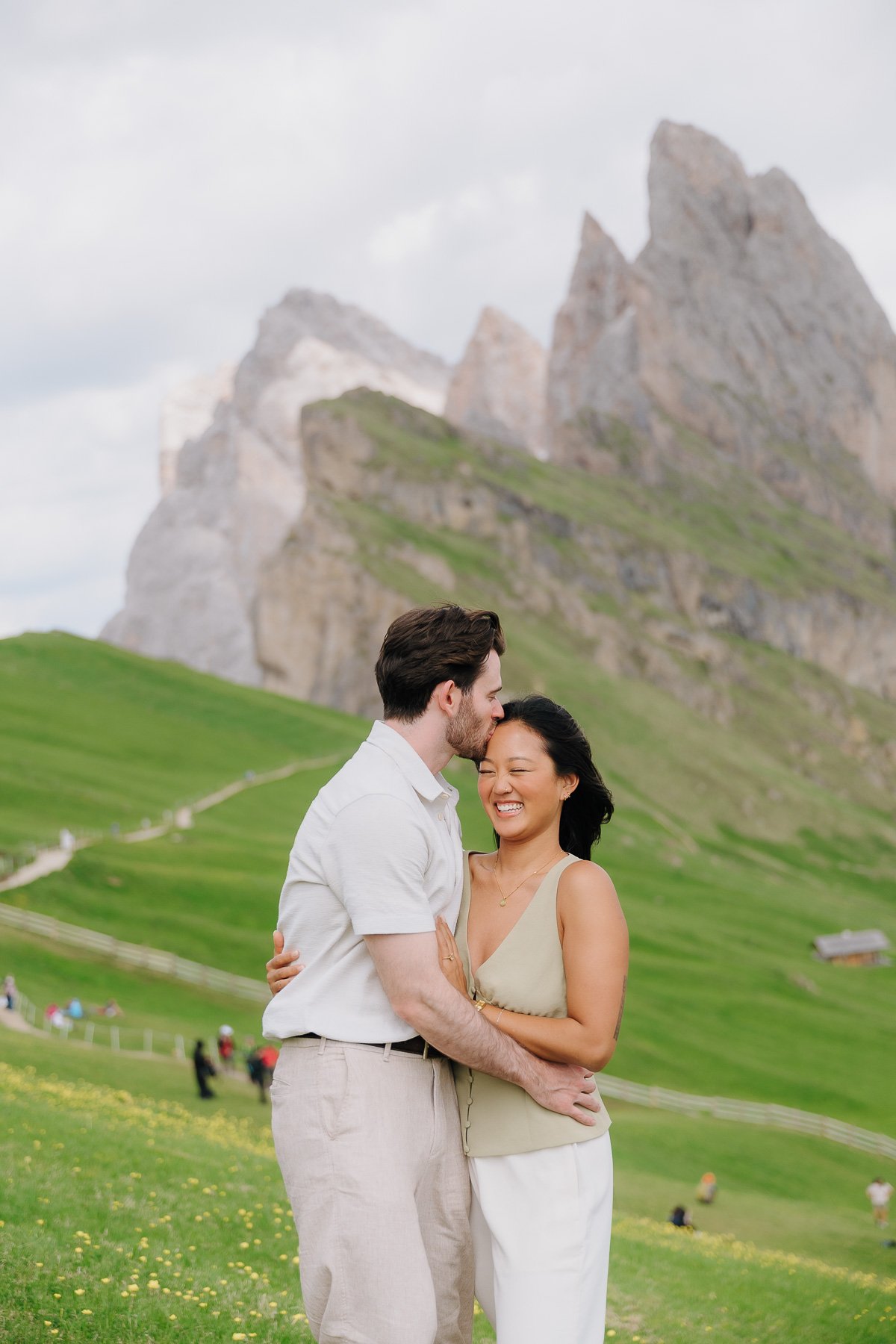 A happy couple embracing in a mountainous field, with rocky peaks and green hills in the background and other people in the distance.
