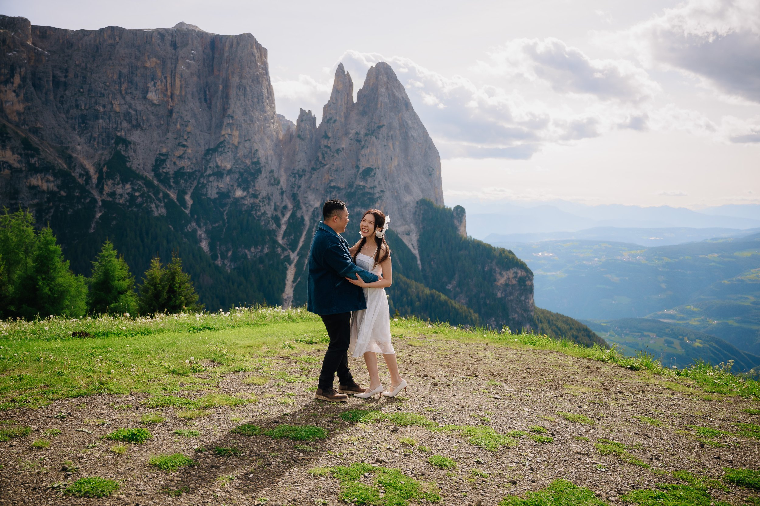 A couple, a man and a woman, standing on a grassy hill with mountains in the background. They are smiling and looking at each other, holding hands.