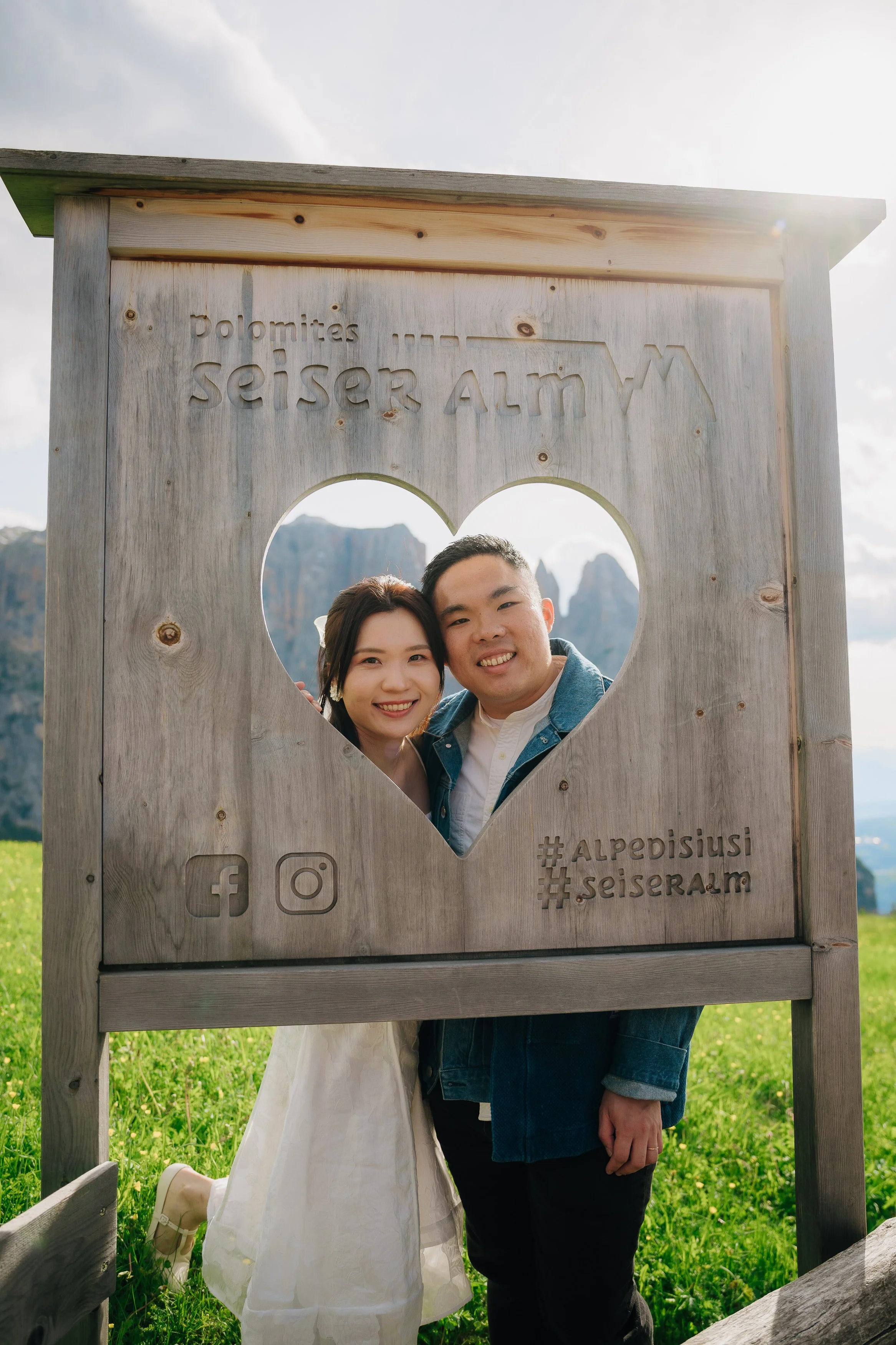 A young couple smiling and posing behind a wooden heart-shaped frame with engraved text, in a lush green outdoor setting with mountains in the background, during daytime.