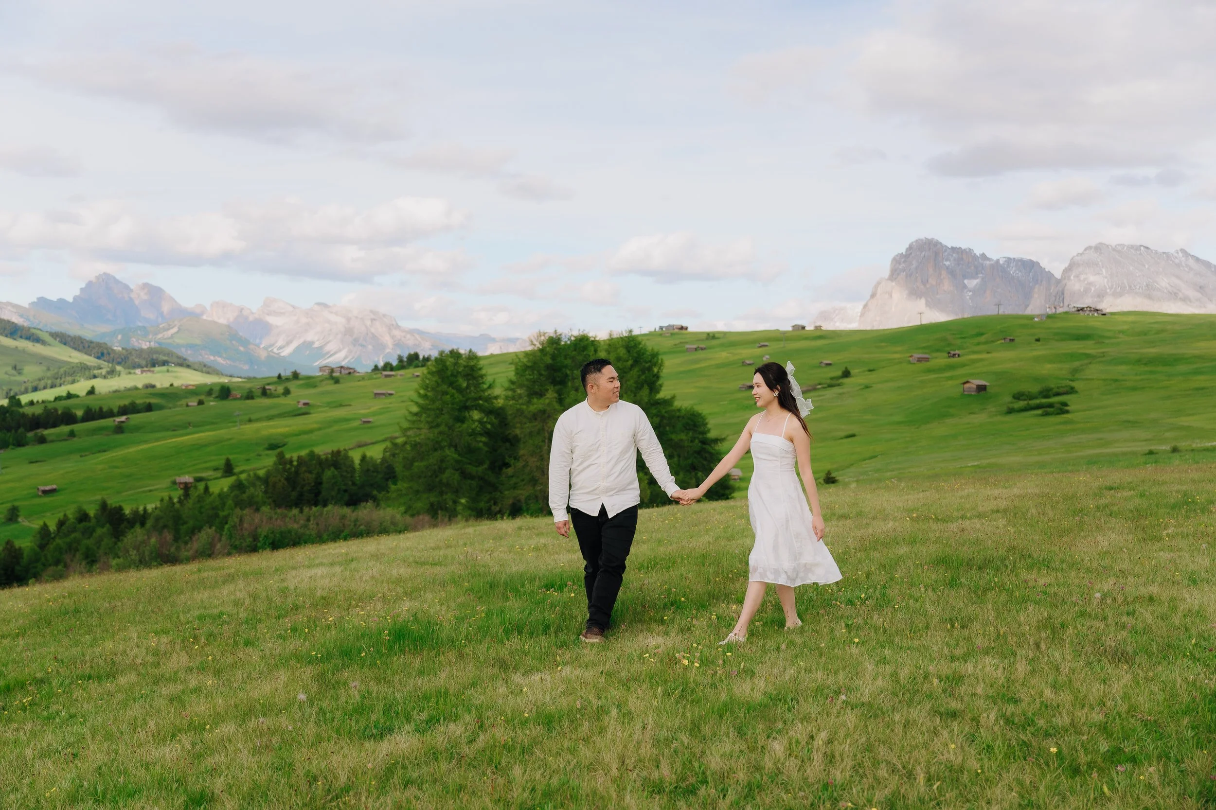 A couple holding hands and walking in a green meadow in front of mountains, with a cloudy sky overhead.