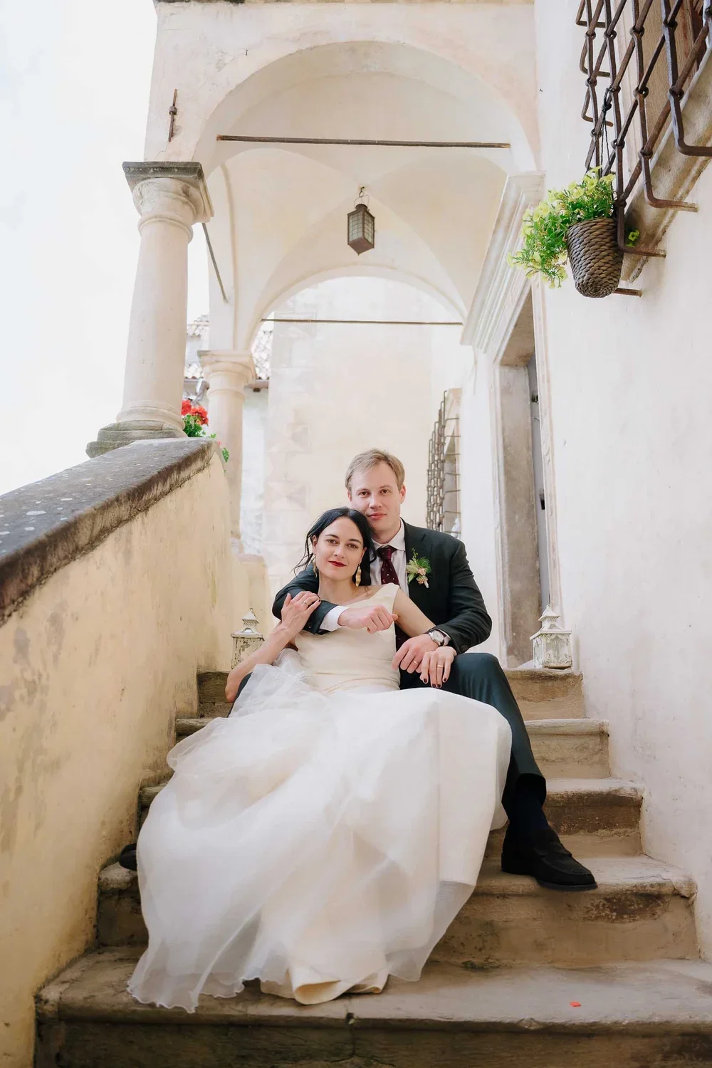 A newlywed couple sitting on stone stairs between light-colored walls with decorative columns and arches, in an outdoor setting with hanging plants and lanterns.