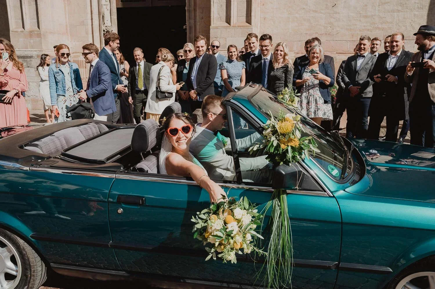 A bride and groom in a teal convertible car decorated with flowers, surrounded by friends and family during a wedding celebration outside a church.