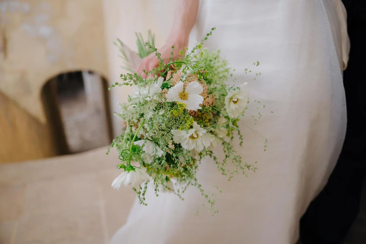 A person holding a bouquet of white and cream-colored flowers with green foliage, with part of a white dress visible.