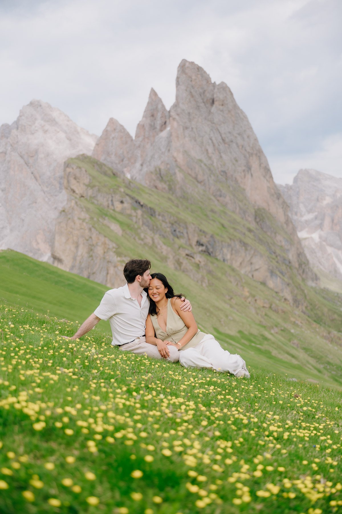 A happy couple sitting on a grassy field with small yellow flowers, with mountains in the background, embracing and smiling.
