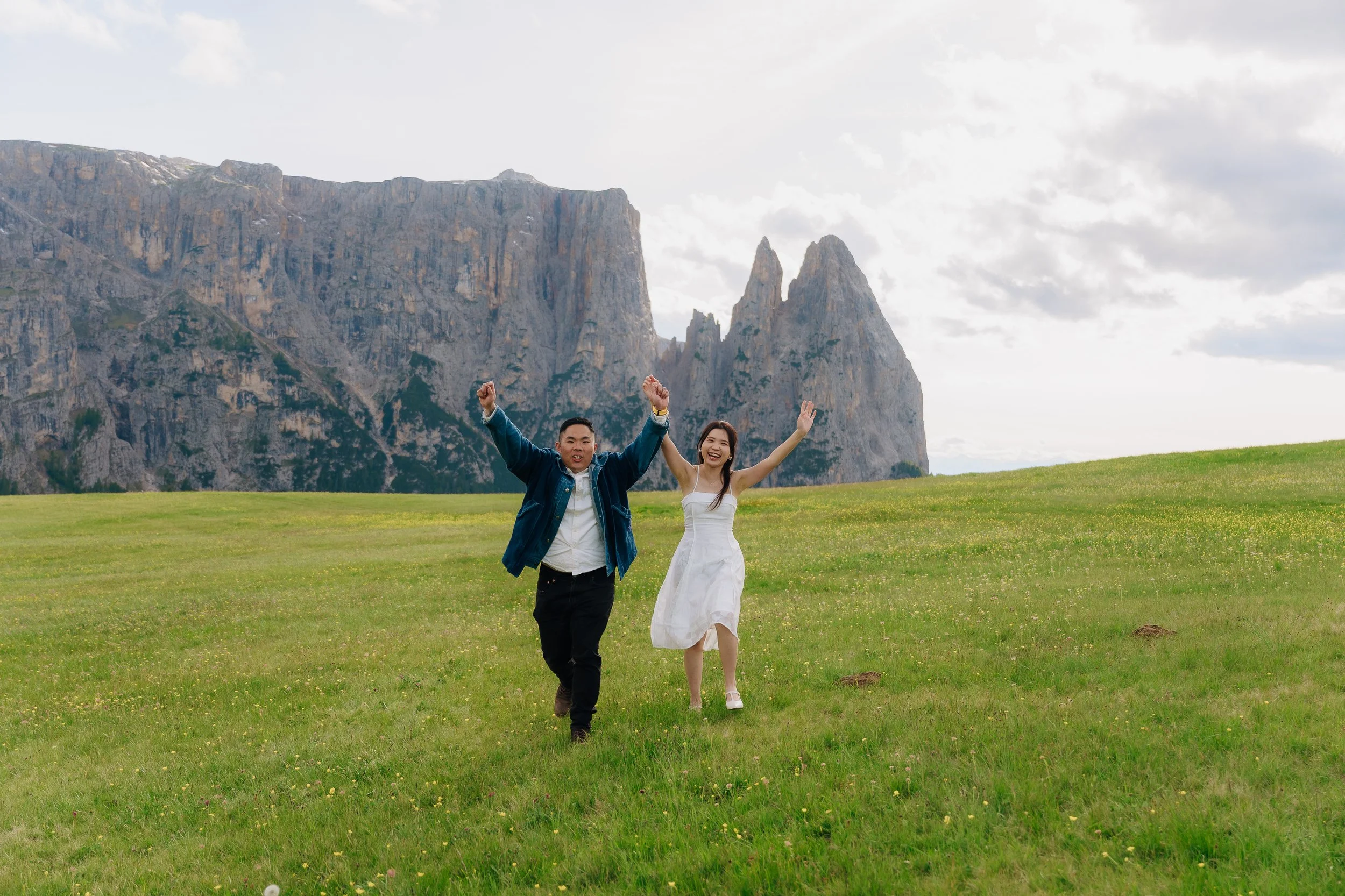 A happy couple running on a grassy field with mountains in the background, the man raising his arms in celebration, and the woman smiling with arms raised.