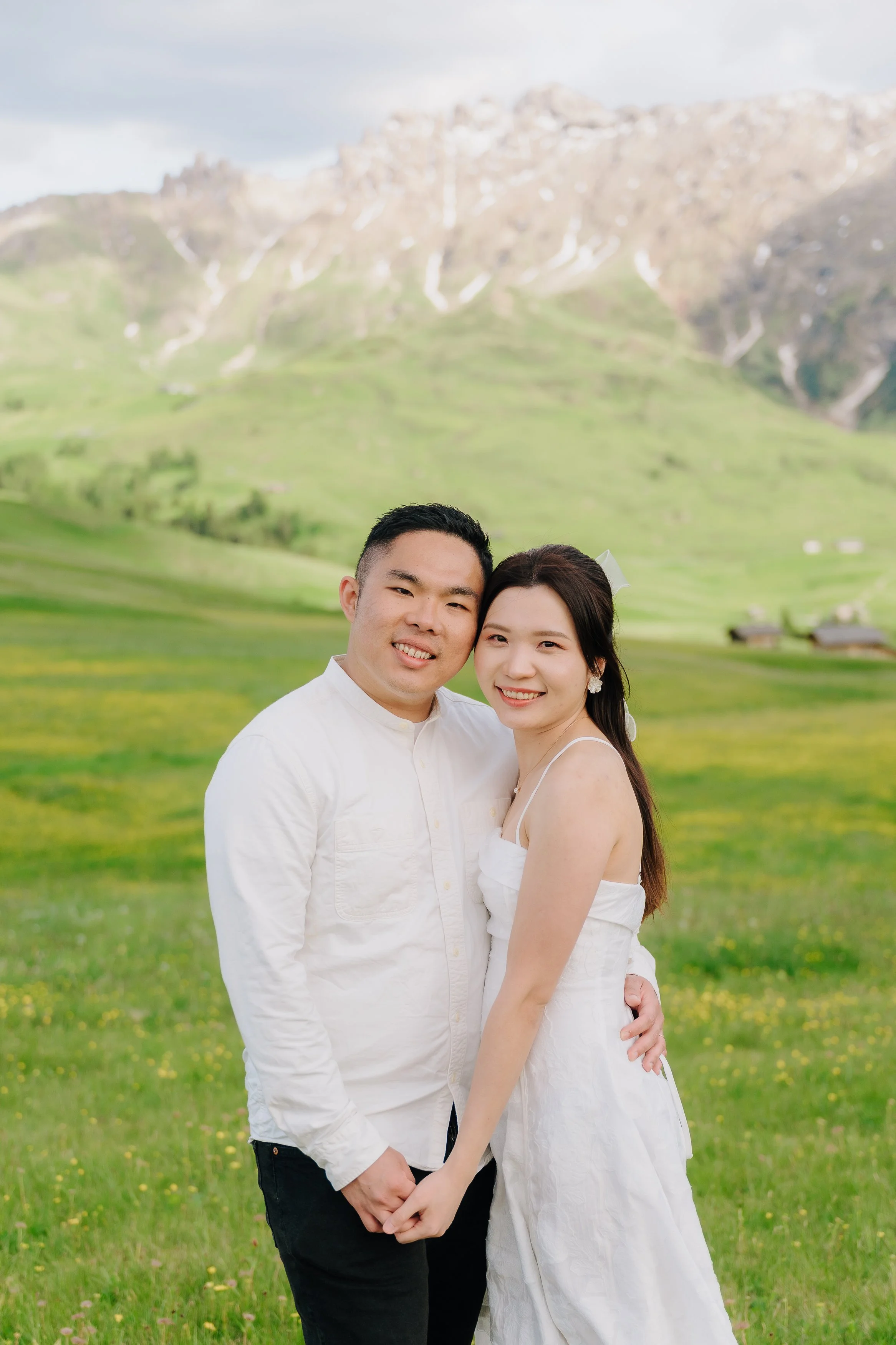 A smiling couple posing outdoors in a lush green field with mountains and a cloudy sky in the background.