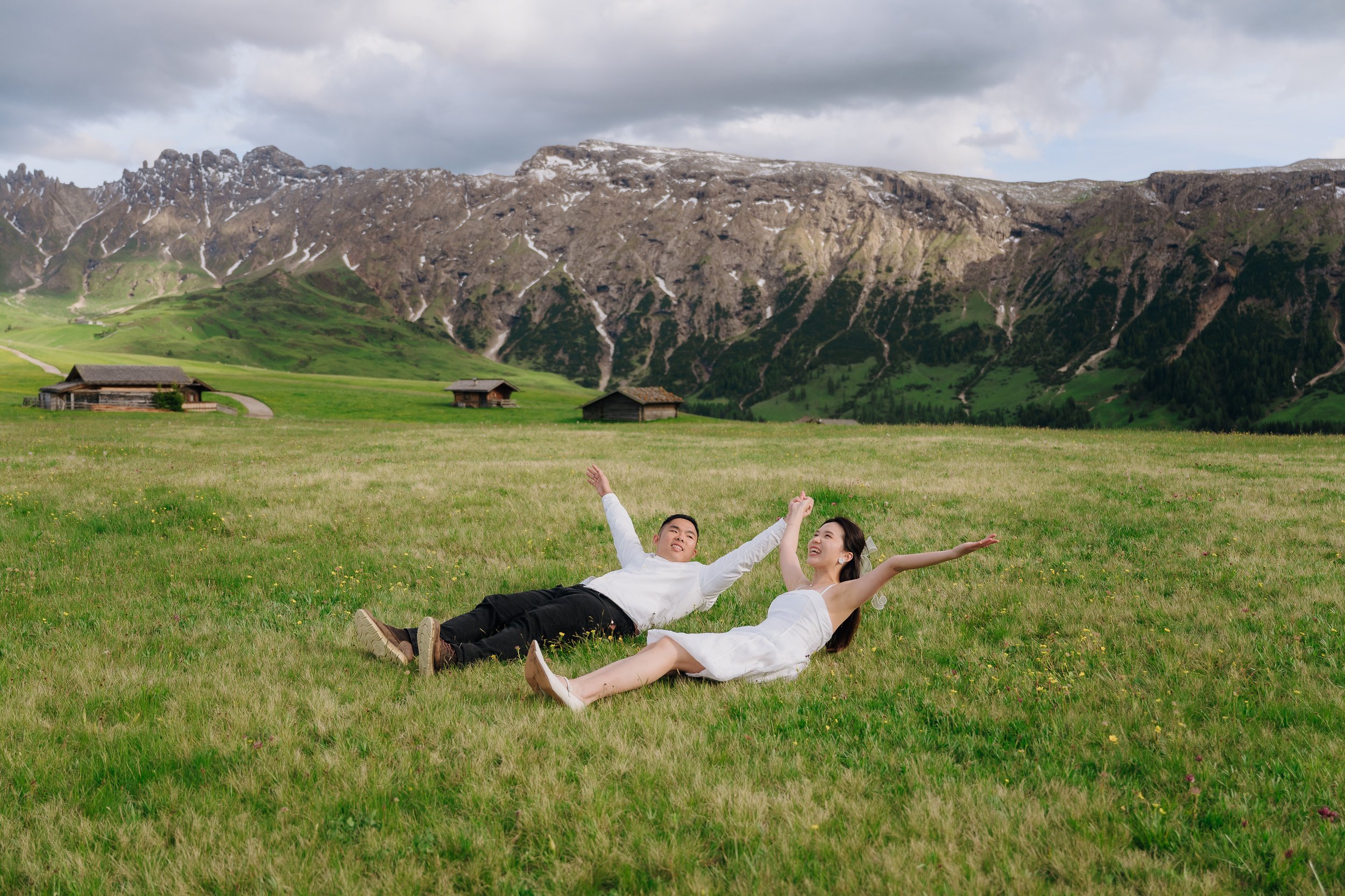Two people, a man and a woman, lying on a grassy field with their arms outstretched, smiling, with a backdrop of mountains and small wooden houses.