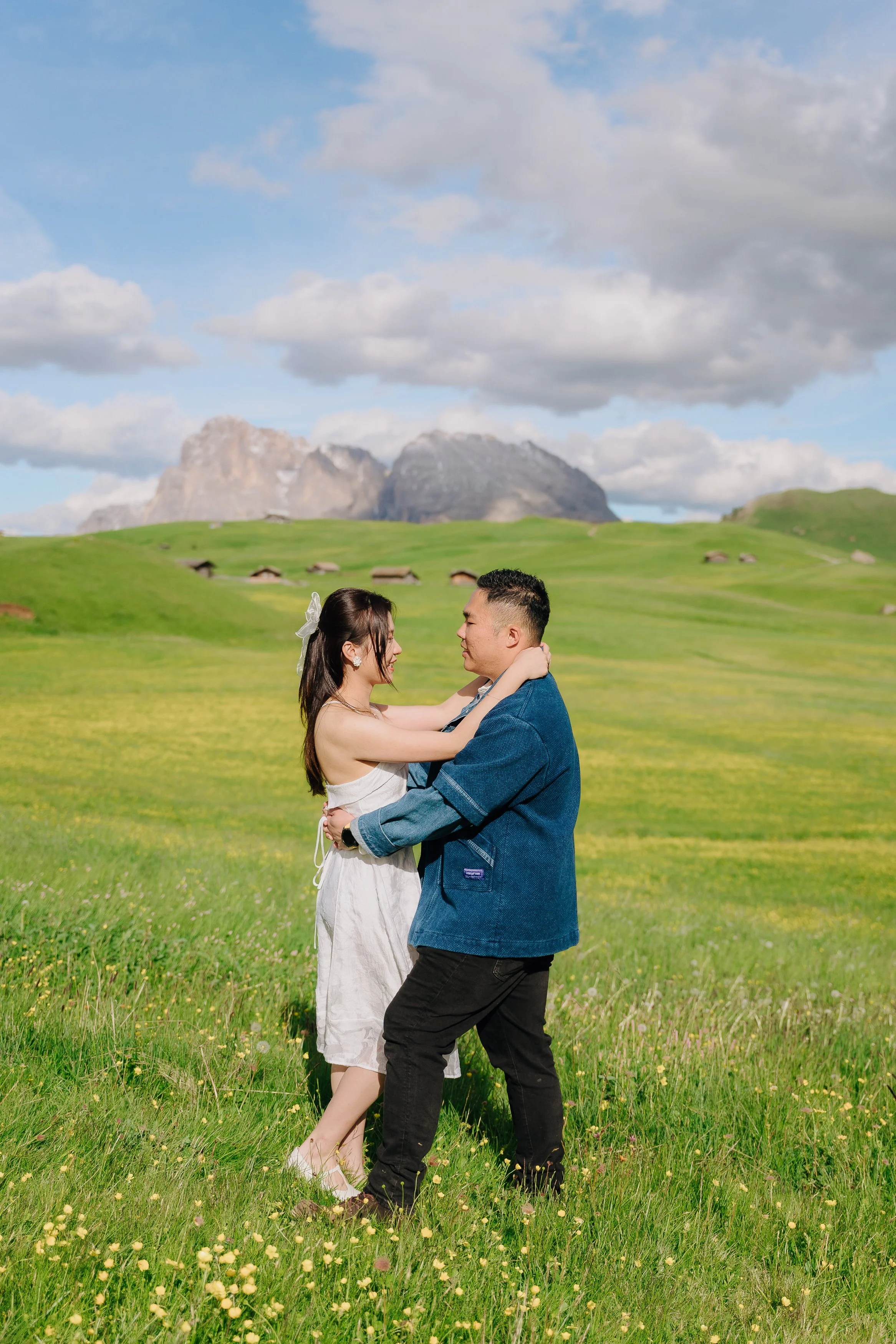 A couple embracing and looking into each other's eyes in a green field with mountains in the background and a cloudy sky.