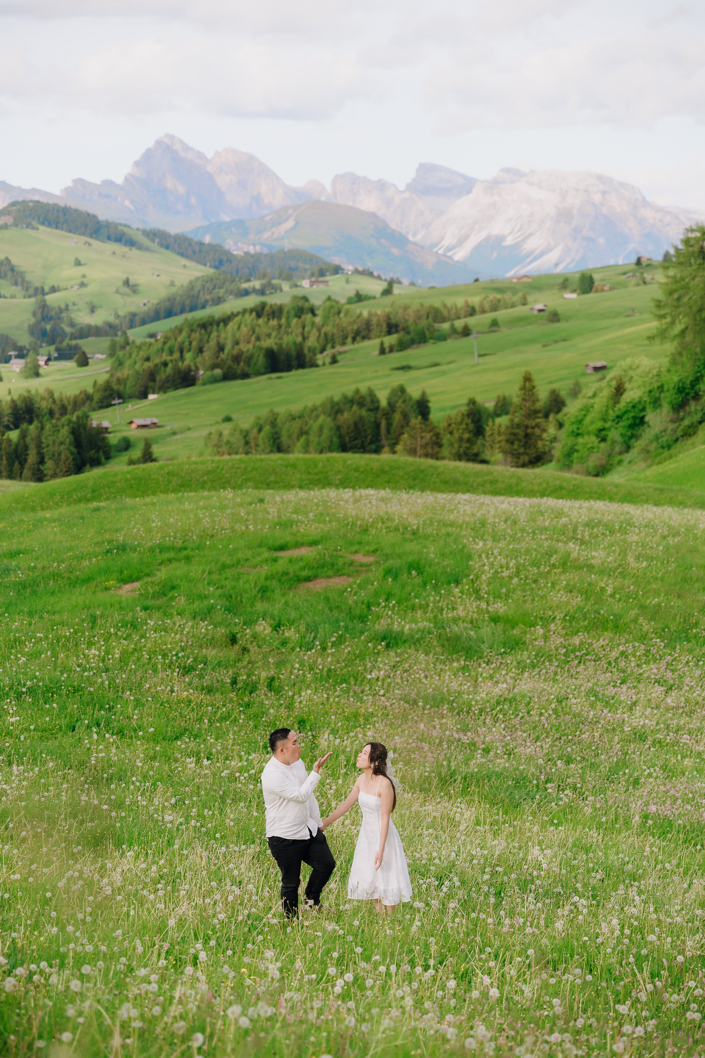 A couple dressed in wedding attire standing in a green field of wildflowers, with rolling hills, scattered houses, and mountains in the background under a partly cloudy sky.