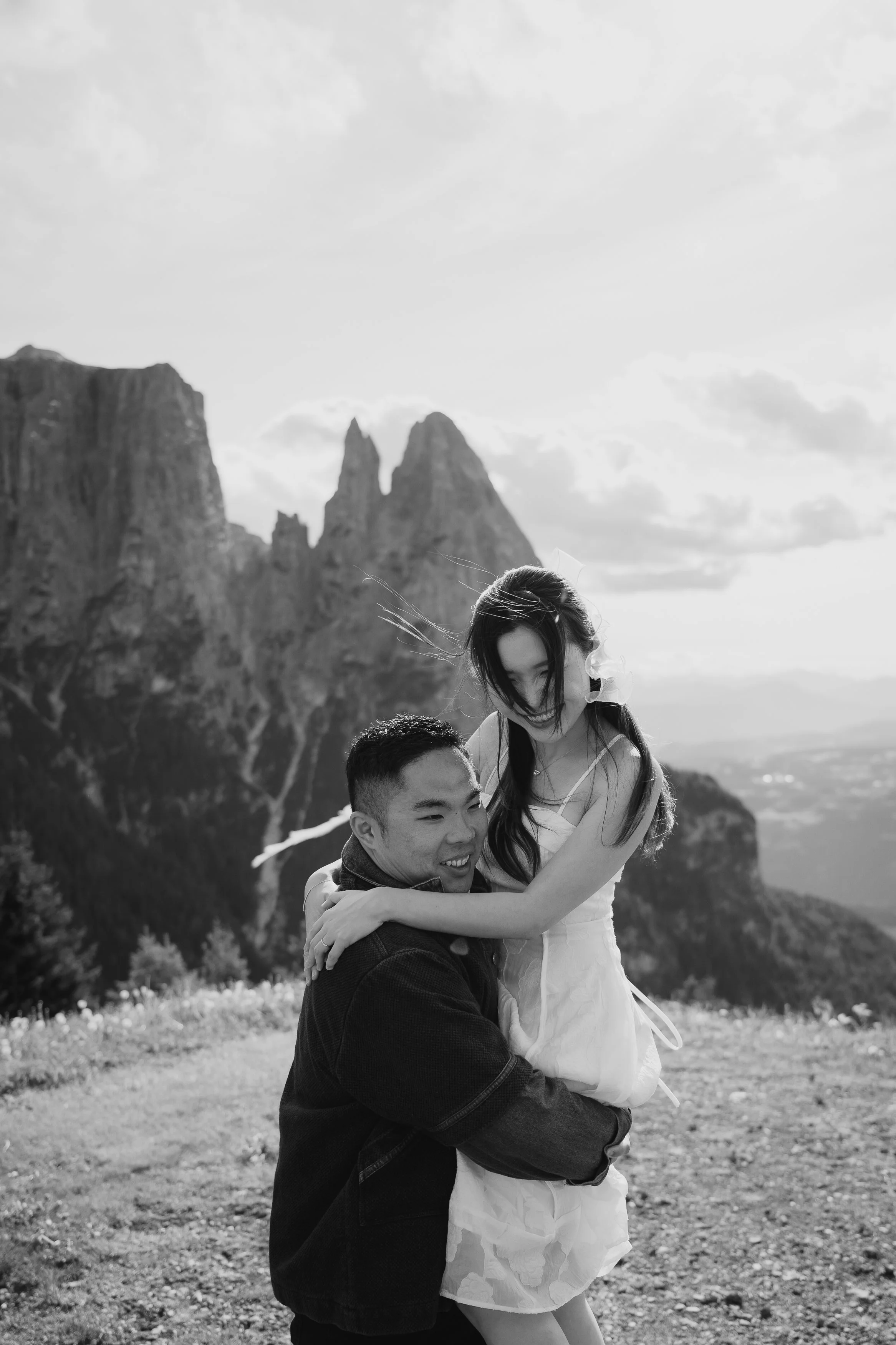 A black-and-white photo of a couple outdoors with a mountain range in the background. The man is kneeling, holding the woman in his arms, and they are smiling. The woman is leaning forward, looking down, with her arms around the man's shoulders.