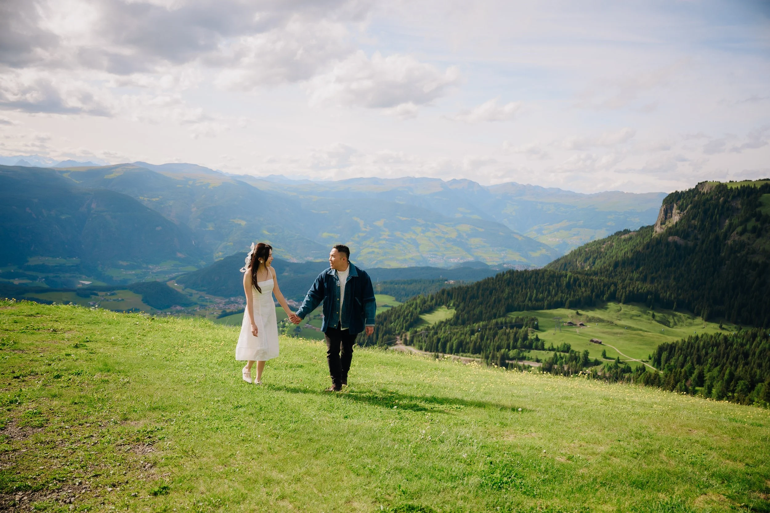A couple holding hands and walking on a grassy hilltop with mountains in the background during daytime.