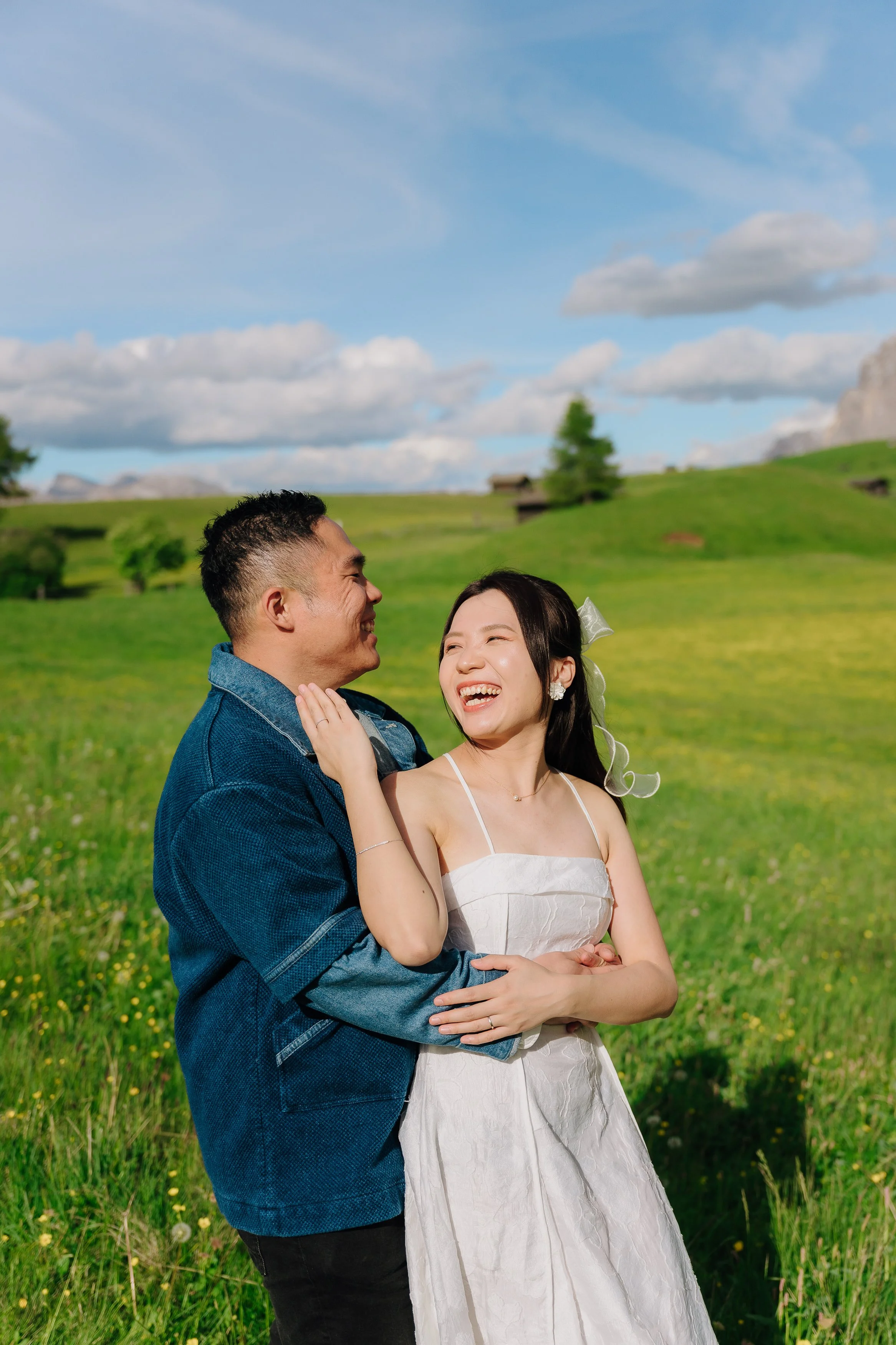 A smiling couple, man in a blue denim jacket and woman in a white dress with a bow in her hair, standing outdoors in a green field with hills, trees, and a partly cloudy blue sky in the background.