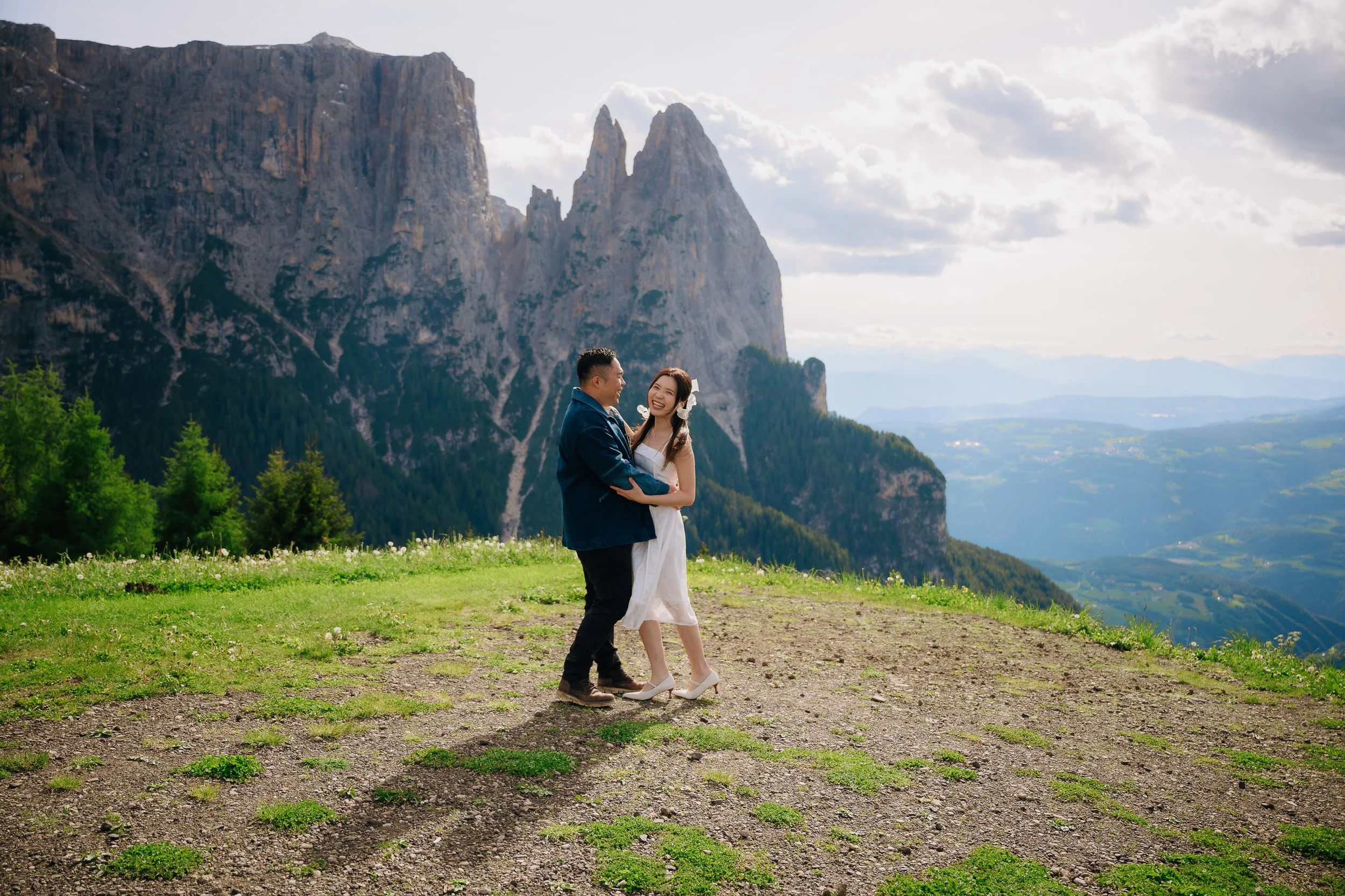 A couple happily dancing in a scenic mountain landscape with lush green trees, distant mountains, and a partly cloudy sky.