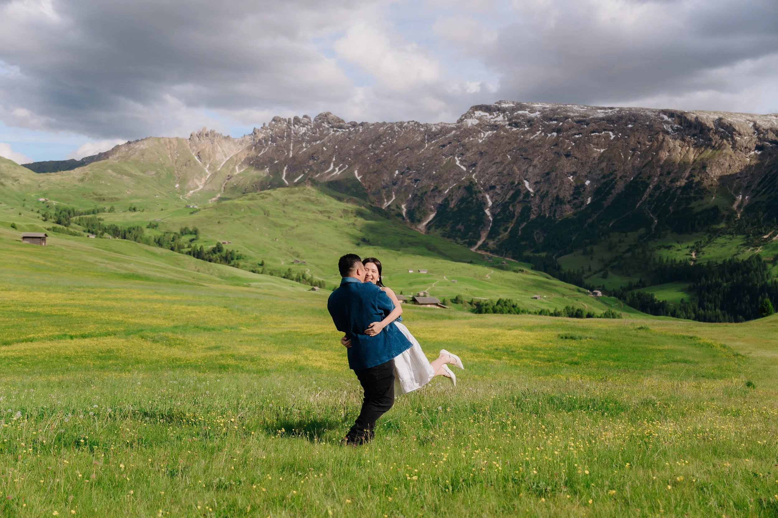 A man in a dark blue jacket holding a smiling woman in a white dress in a green field with mountains in the background.