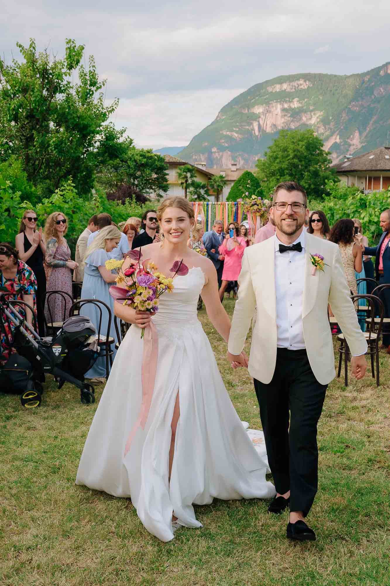 A bride and groom walking hand in hand at an outdoor wedding ceremony. The bride is holding a bouquet of flowers and is dressed in a white wedding gown. The groom is wearing a white tuxedo jacket with a black bow tie and black pants. Behind them, guests are dressed in colorful attire, and the setting features lush green trees and mountains in the background.