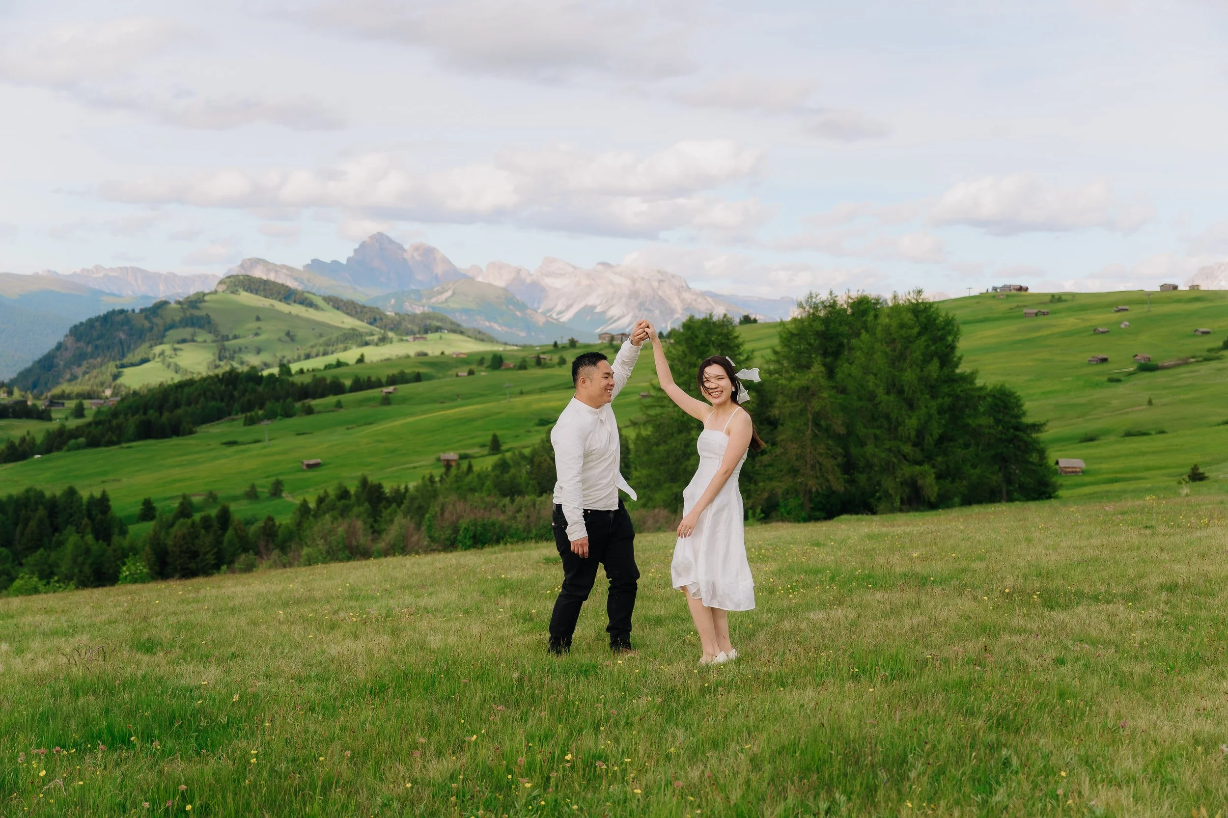 A couple dancing in a green field with mountains and a cloudy sky in the background.