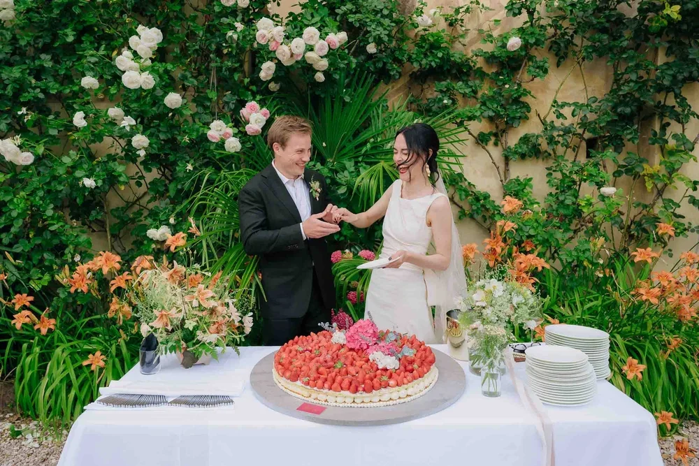 A bride and groom exchanging rings over a wedding cake at an outdoor reception, surrounded by flowers and greenery.