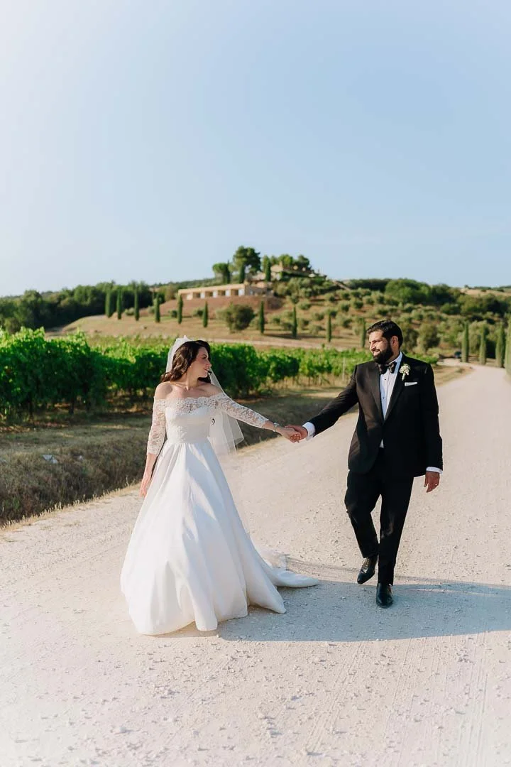A bride and groom holding hands and walking outdoors on a dirt road, with a backdrop of greenery, trees, and a hill with a structure on top. In tuscany Italy