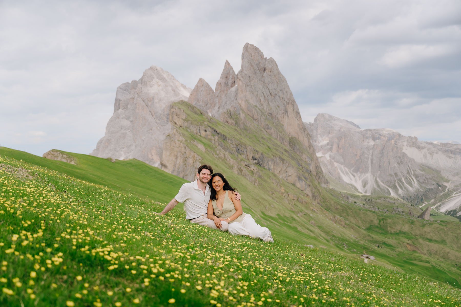 A couple sitting and smiling on a grassy hillside covered with small yellow flowers, with green mountains and a cloudy sky in the background.
