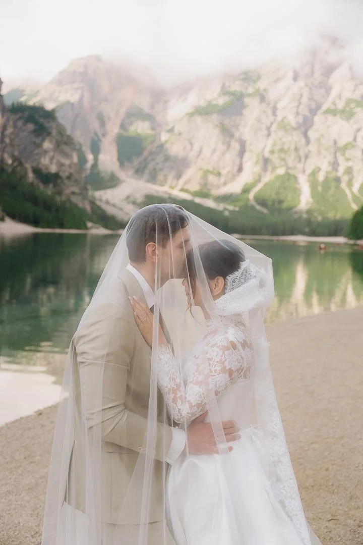 Bride and groom kissing with a veil over their faces beside a lake and mountain landscape. at Braies Lake Dolomites