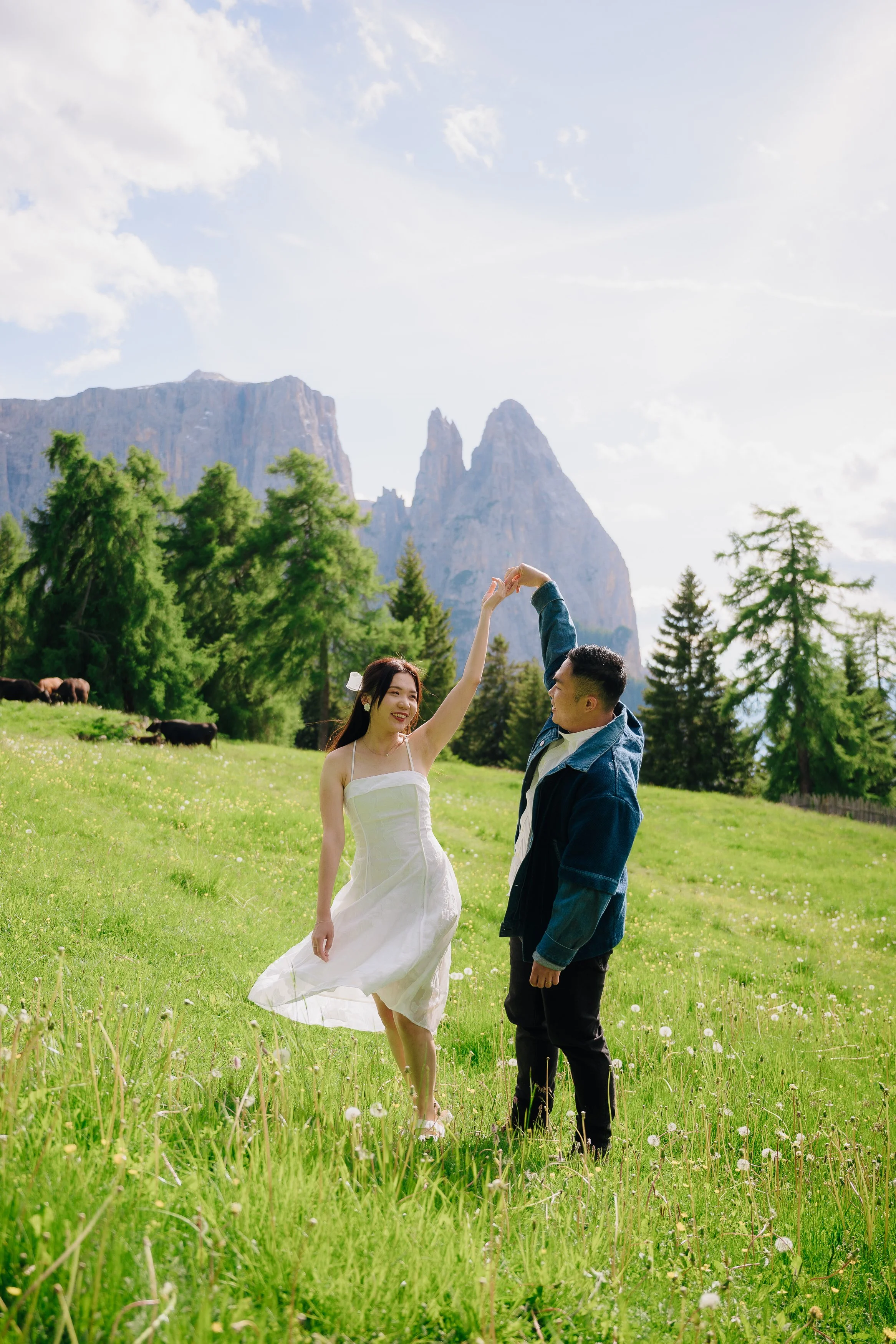 A couple dancing in a green meadow with mountains and trees in the background, under a partly cloudy sky.