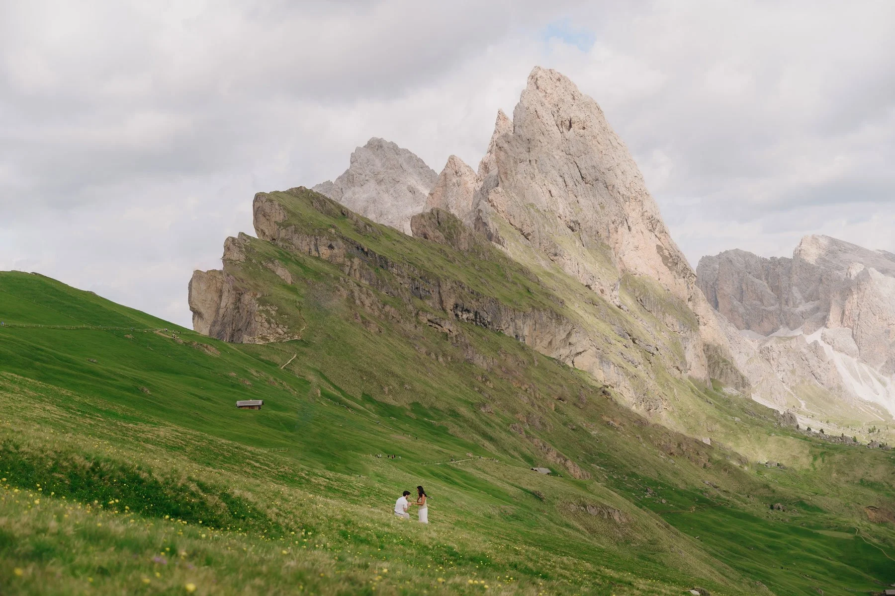 A mountain landscape with rocky peaks and green grassy slopes. Two people are in the foreground, kneeling and facing each other, on a grassy hillside.