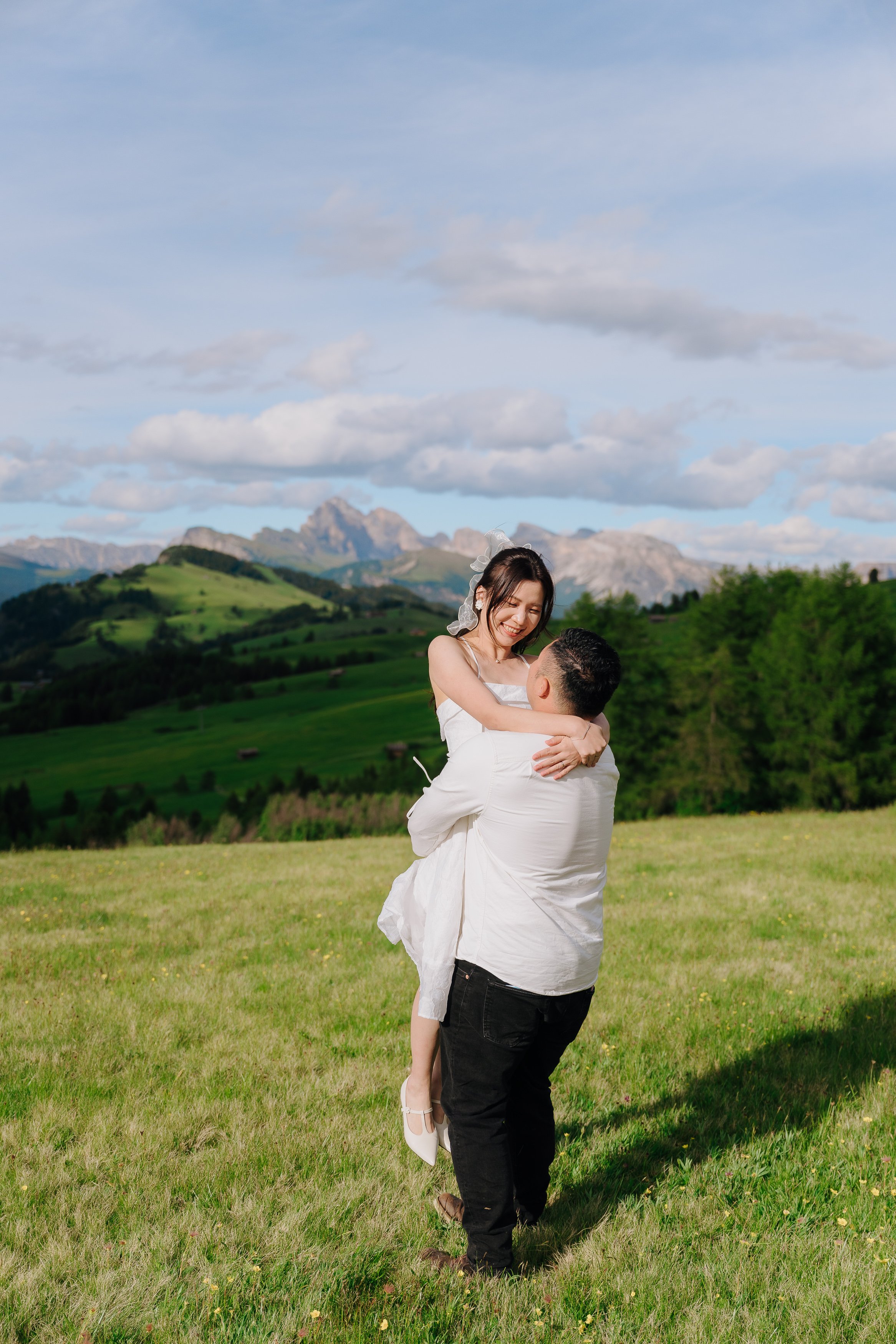 A couple outdoors in a grassy field with rolling hills and mountains in the background. The man is lifting the woman, and they are smiling at each other, dressed in white.