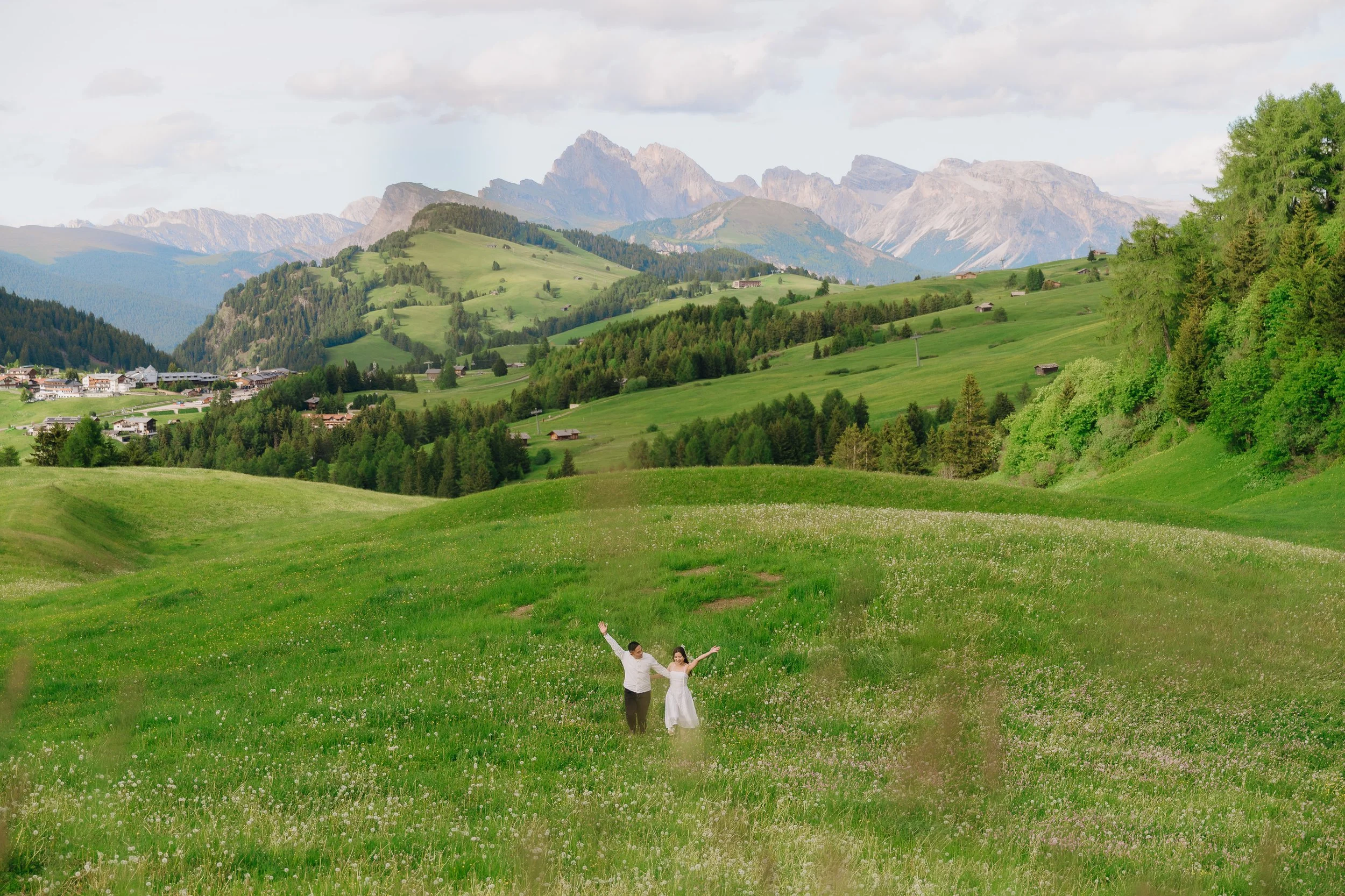 A couple in wedding attire standing in a lush green meadow with rolling hills and mountains in the background.