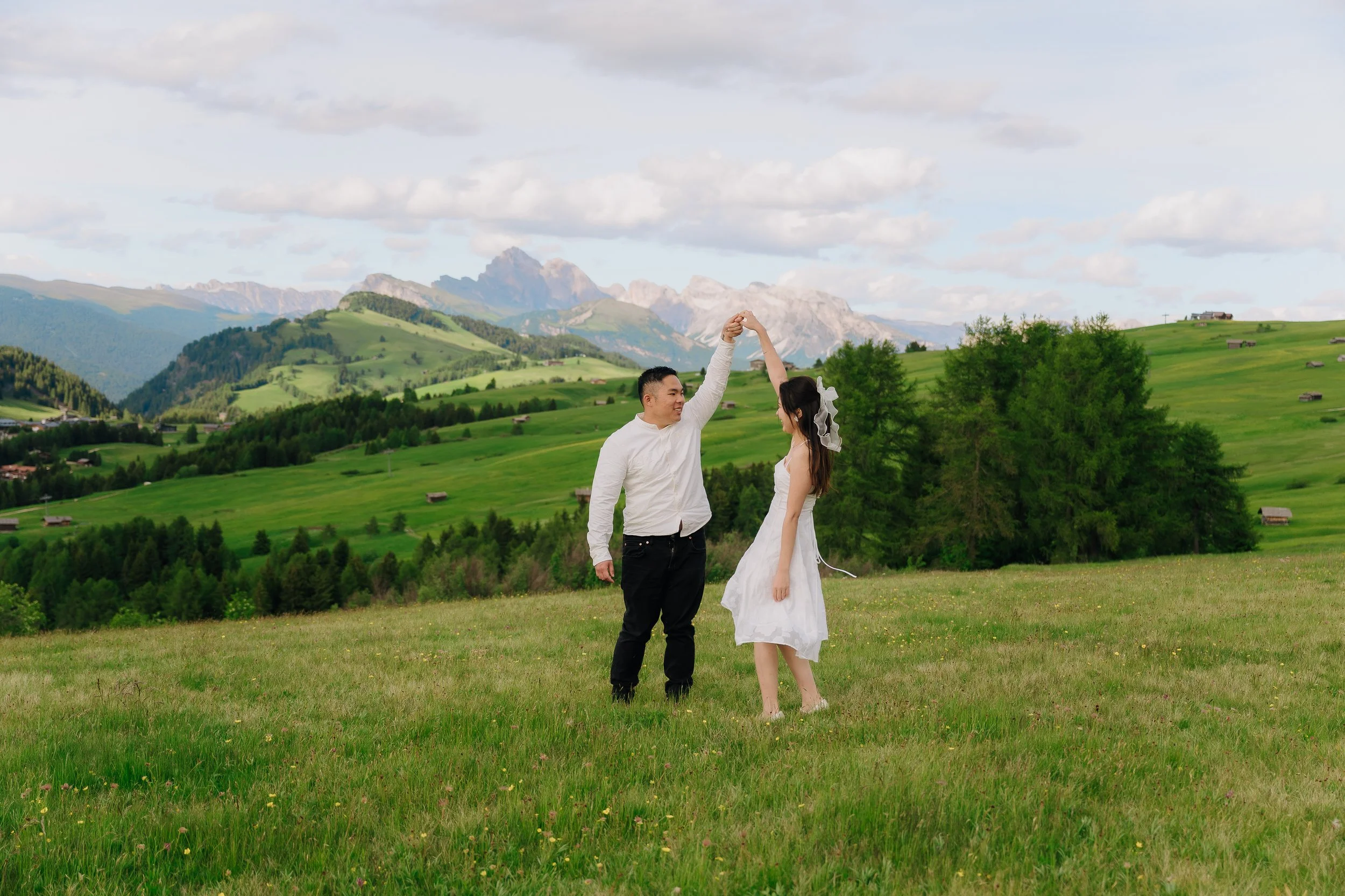A couple dancing in a grassy field with rolling green hills and mountains in the background, under a partly cloudy sky.