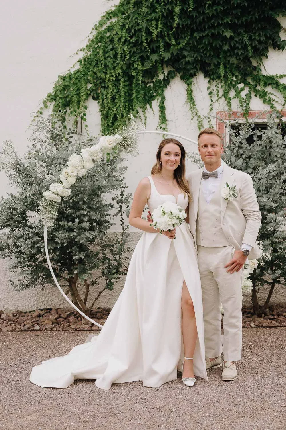 A bride and groom in wedding attire posing outdoors in front of a decorated backdrop with green foliage and a circular floral arrangement, both smiling.