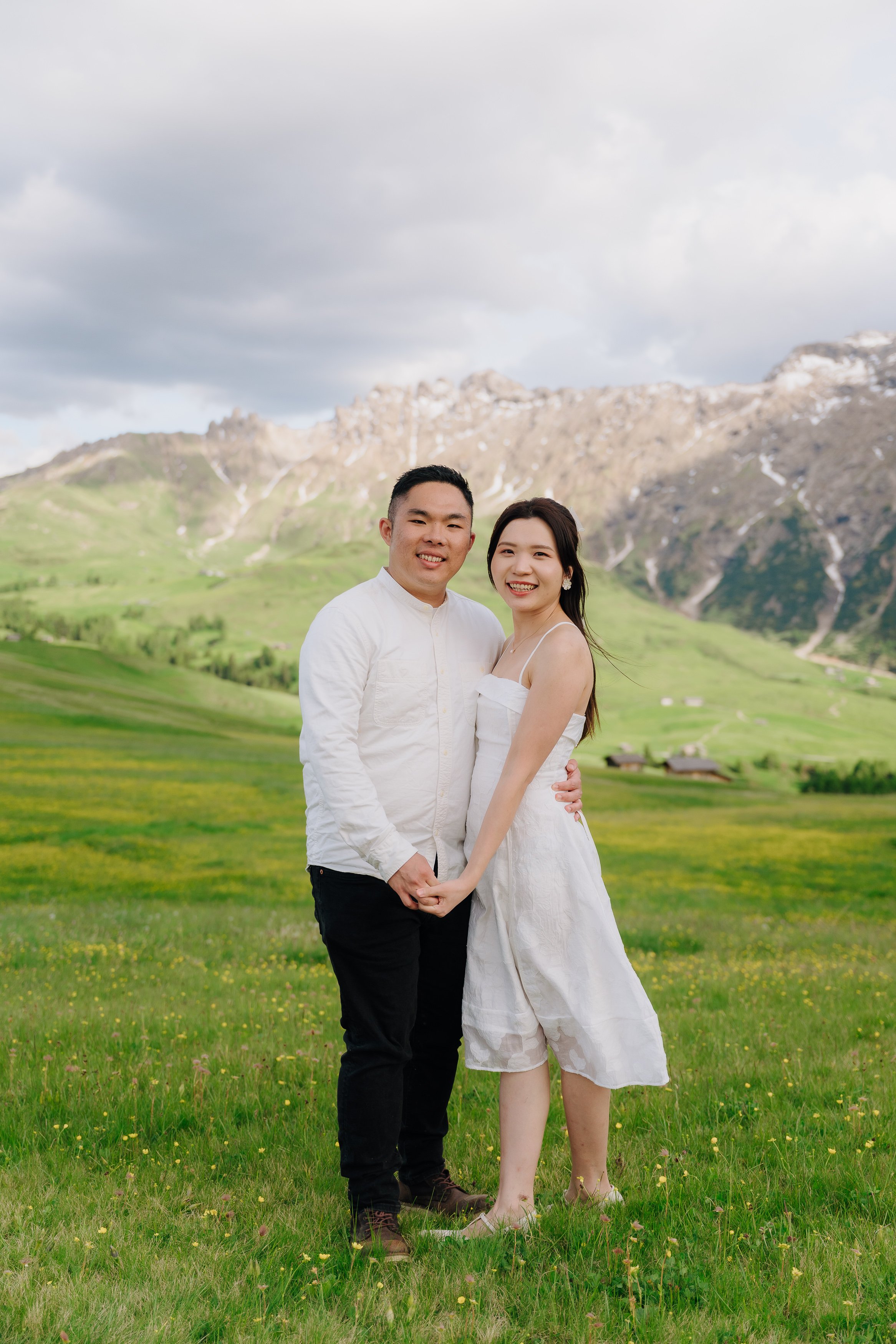 A young couple standing closely together on a grassy field with yellow flowers, holding hands and smiling at the camera. Behind them, there are green rolling hills, small houses, and mountains with patches of snow under a cloudy sky.