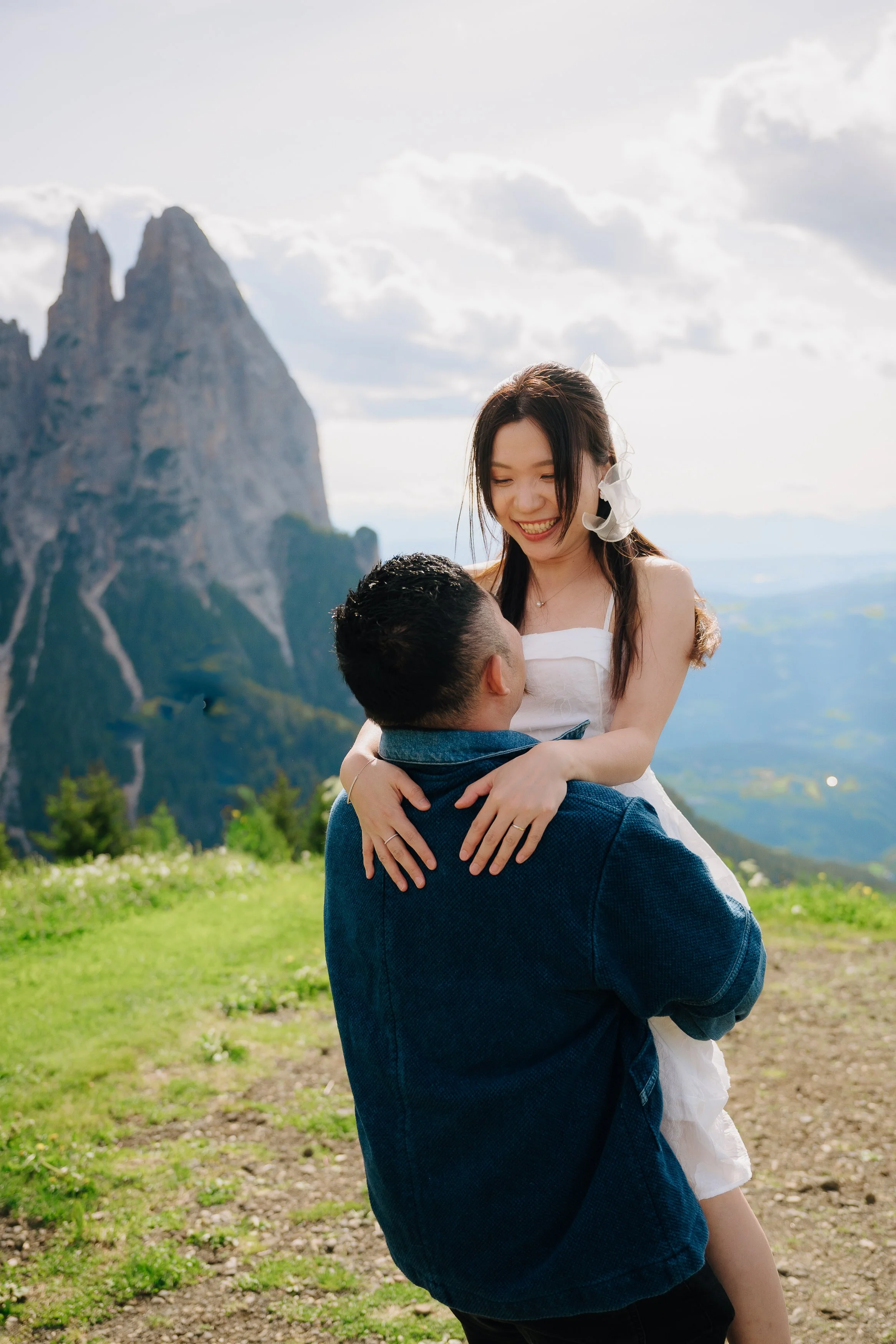 A couple embracing outdoors with a mountainous landscape in the background, the woman smiling with a white dress and a large white flower in her hair, the man wearing a suit. The background features large rocky peaks and a partly cloudy sky.