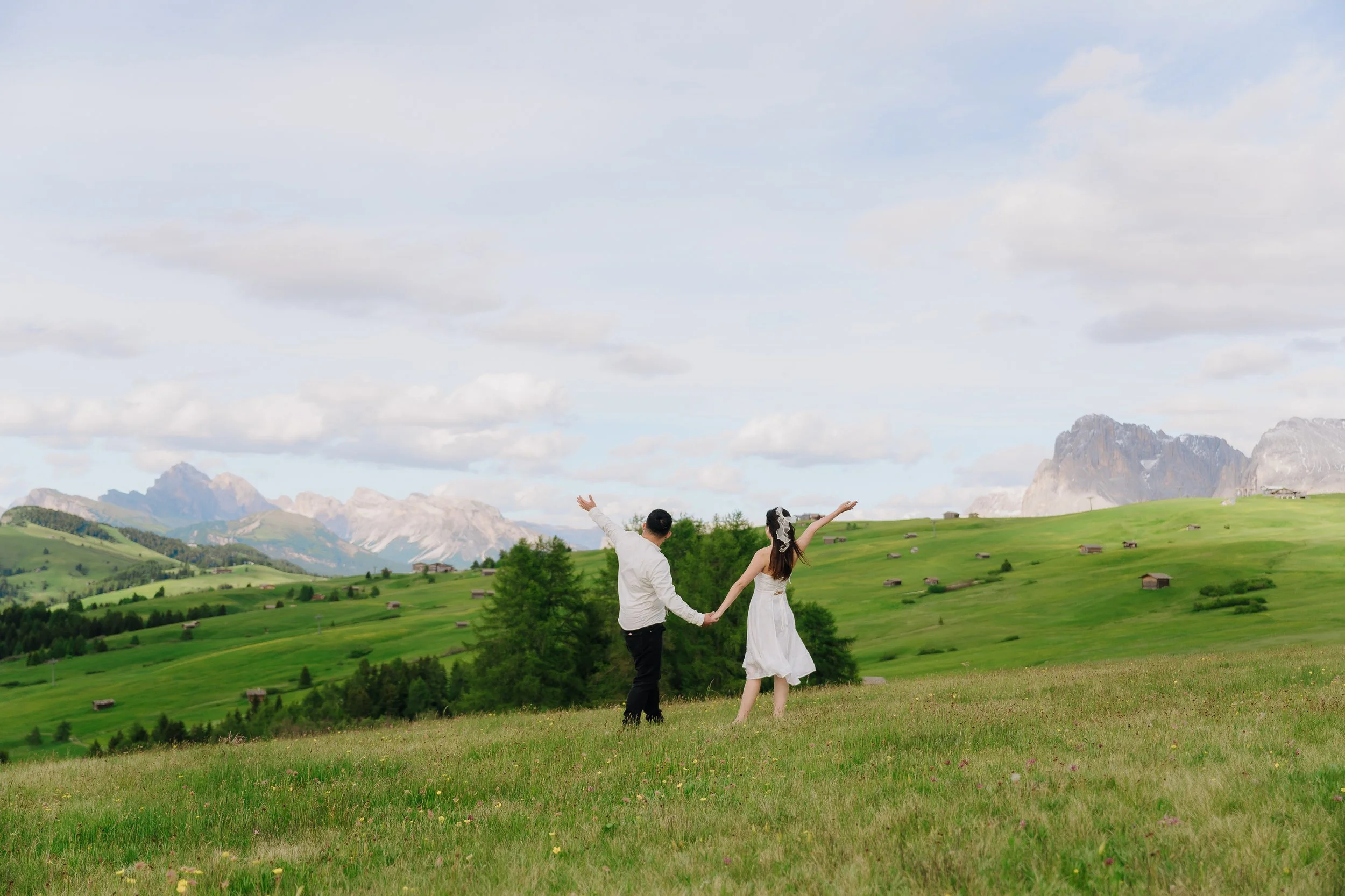 A bride and groom holding hands and dancing in a green field with mountains in the background, under a partly cloudy sky.