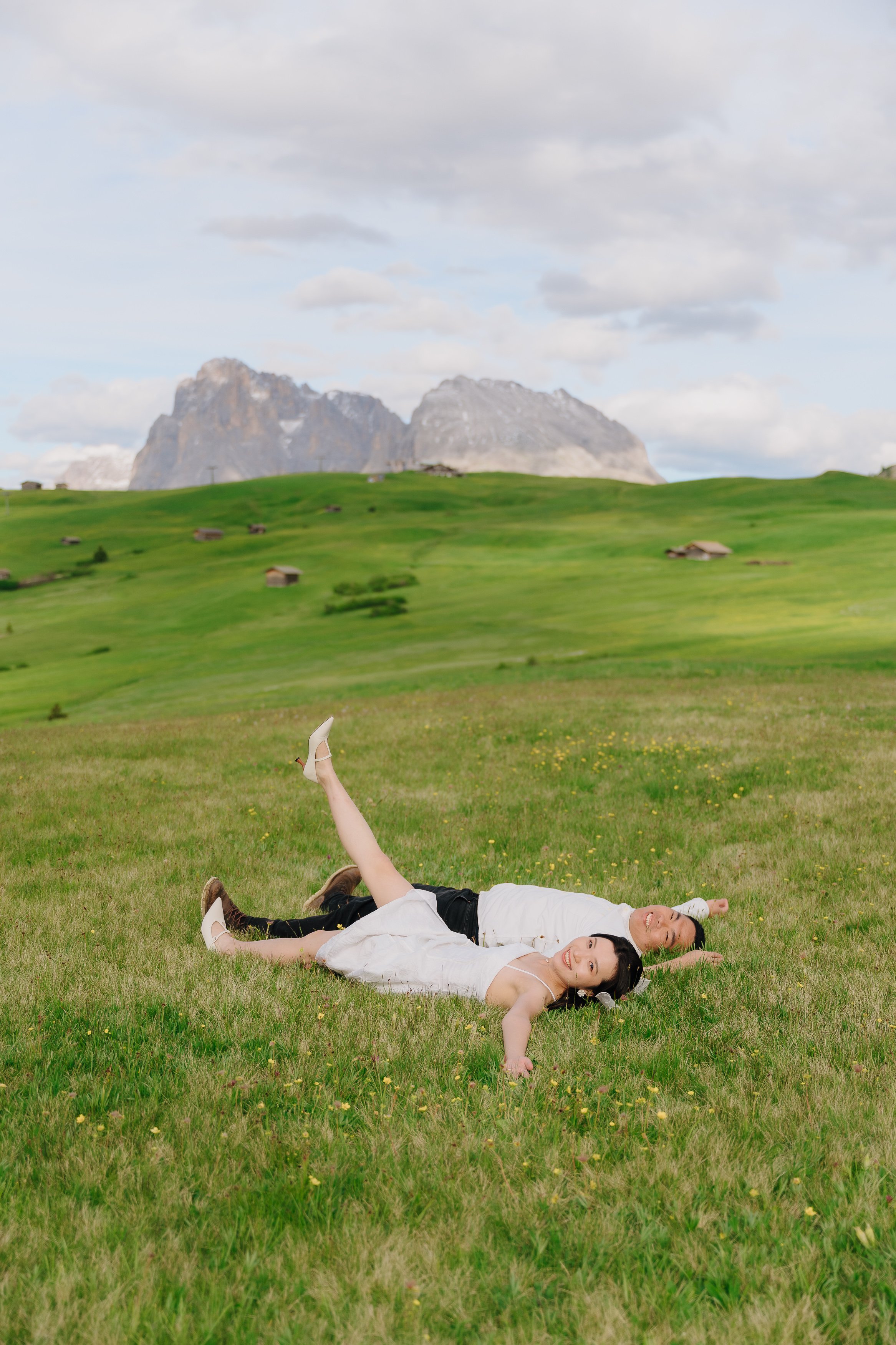 Two women lying on the grass in a field with rolling hills and mountains in the background. They are smiling and appear to be enjoying a sunny day outdoors.