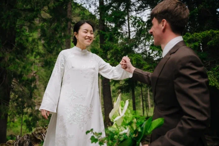 A woman in a white dress and a man in a suit are outdoors among trees, smiling and gently holding hands in a joyful moment.