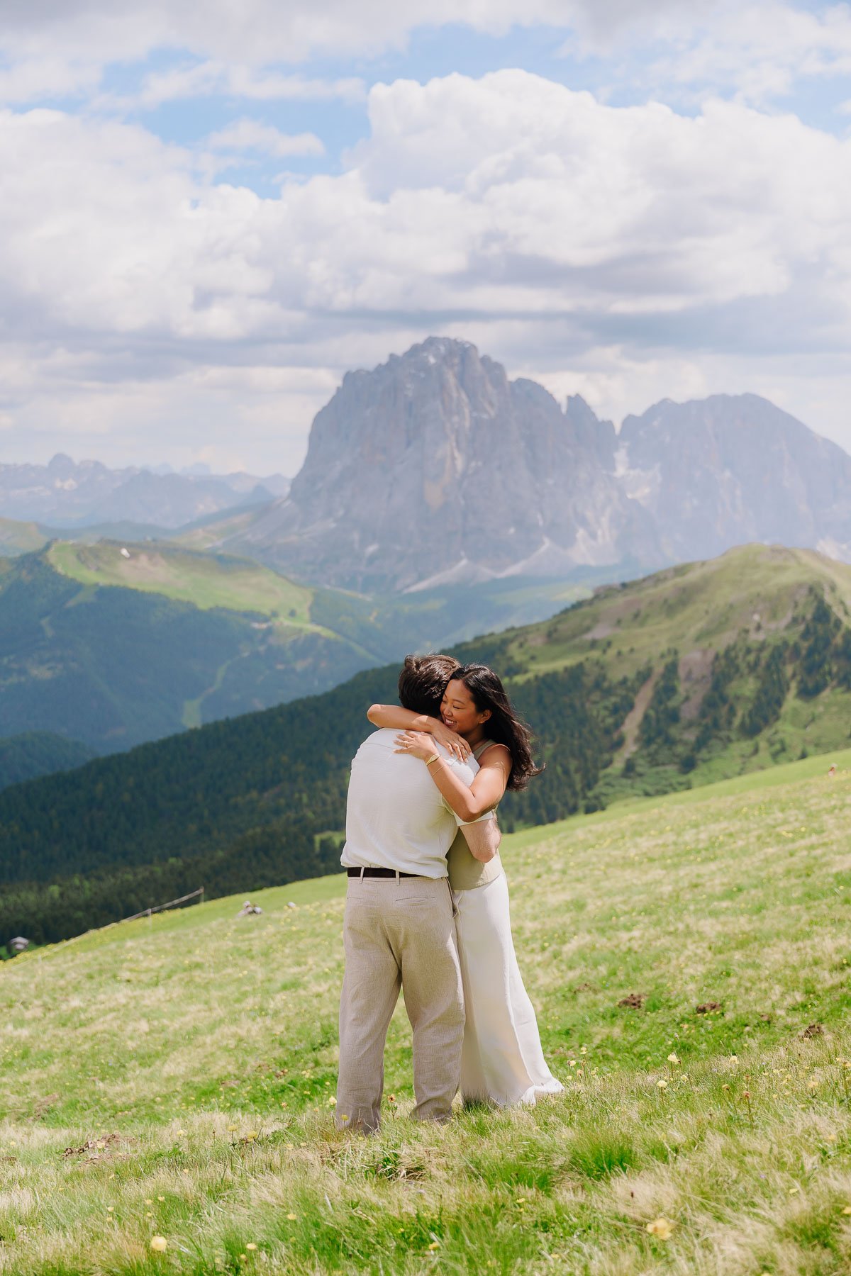 A couple hugging in a grassy field with mountains and a cloudy sky in the background.