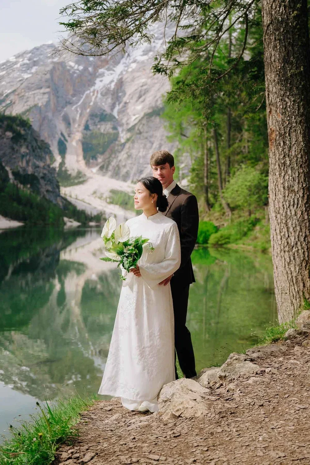 A bride and groom standing on a dirt path by a lake with mountain scenery in the background. The bride is holding a bouquet of white and green flowers, and they are surrounded by trees.