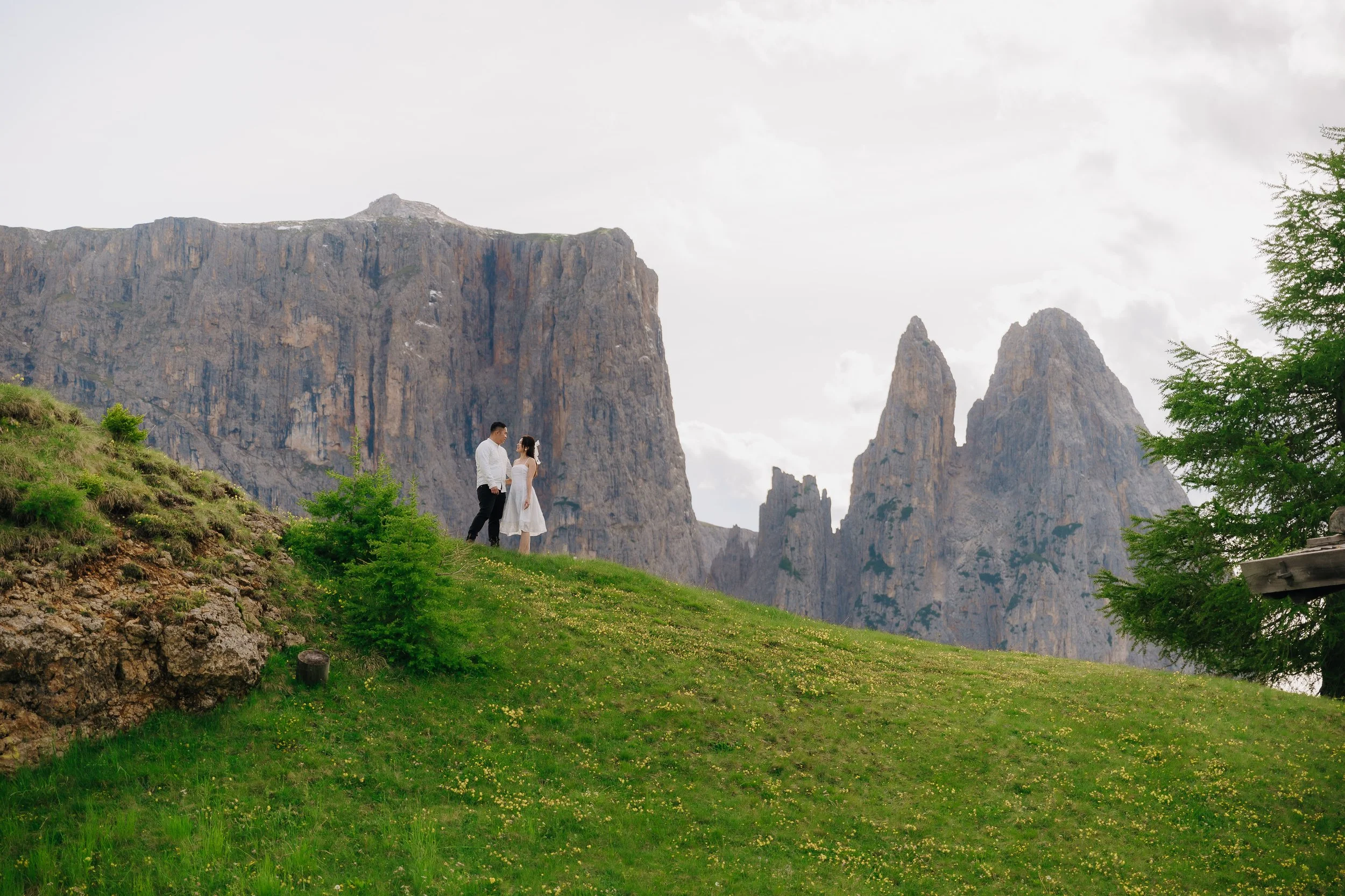 A couple dressed in white standing on a grassy hillside with yellow flowers, facing each other, with tall rocky mountains in the background and a cloudy sky overhead.