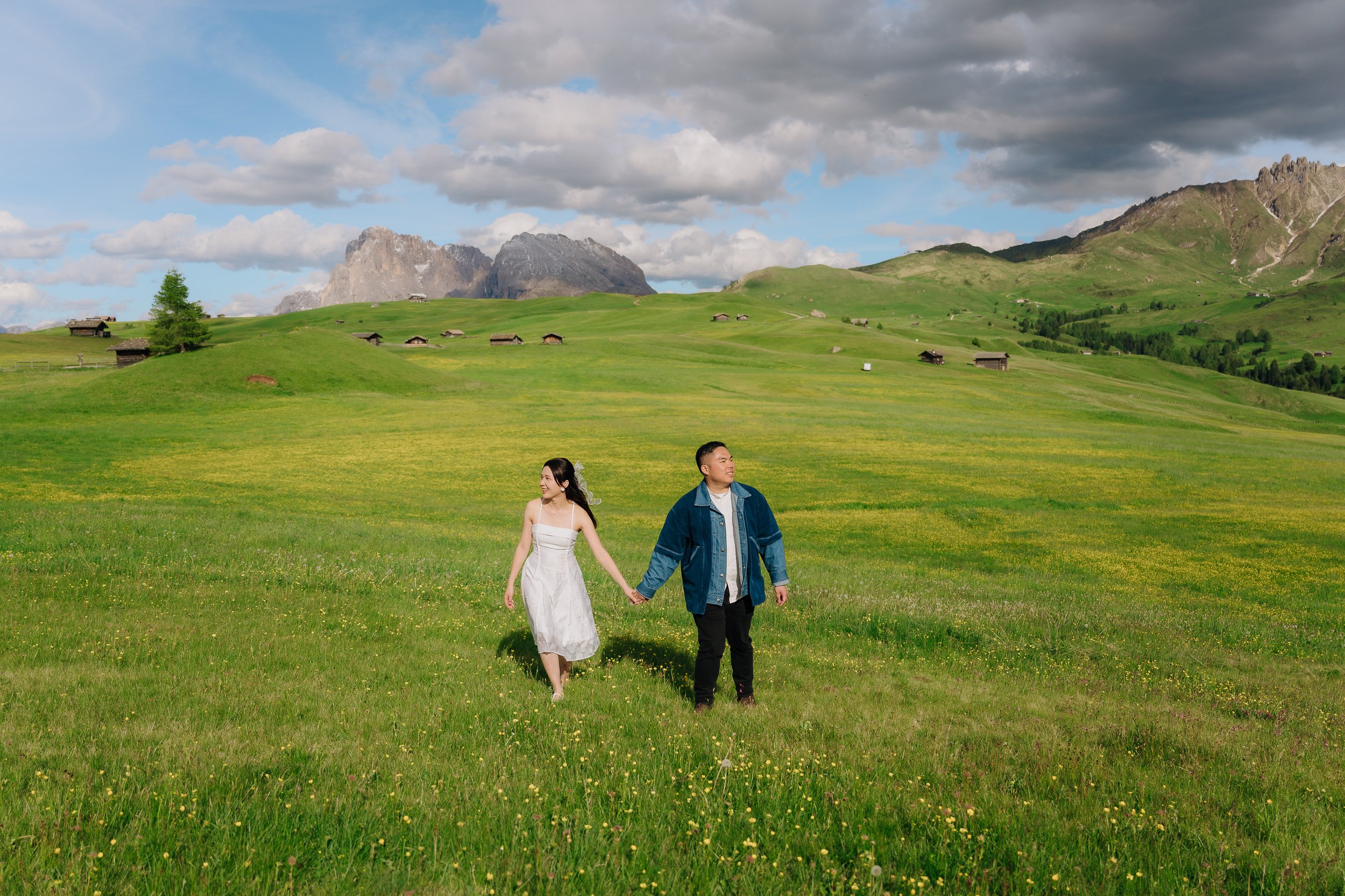 A couple walking hand in hand through a lush green meadow with mountains in the background and a partly cloudy sky overhead.
