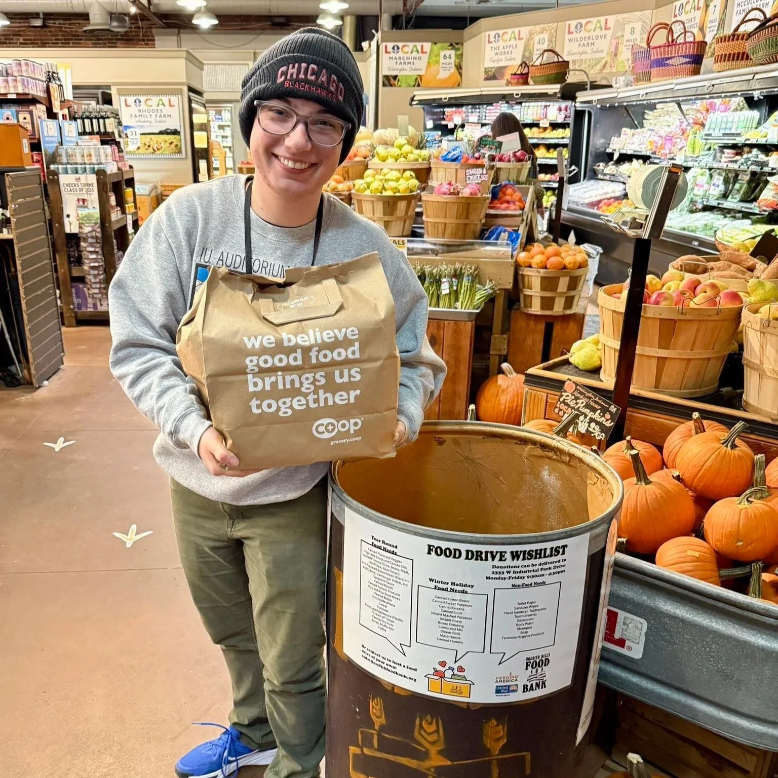 Bloomingfoods team member holding pantry staples bag next to Hoosier Hills Food Bank donation barrel