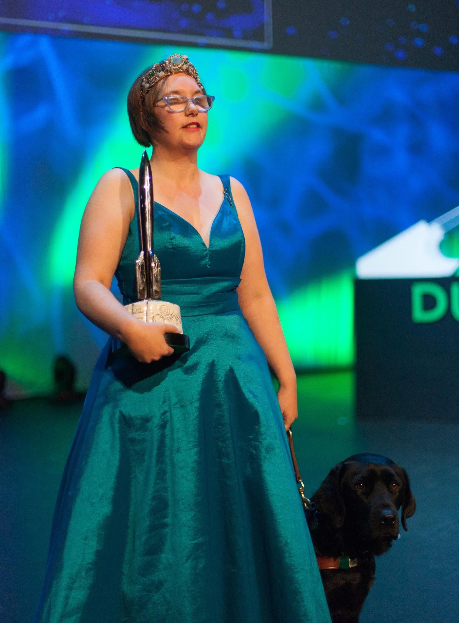 Elsa stands in a teal gown, holding a Hugo Award in the crook of her right arm, and a leash attached to a black Labrador in the left hand. Phjoto by Simon Bubb. Photo used with permission from Simon.