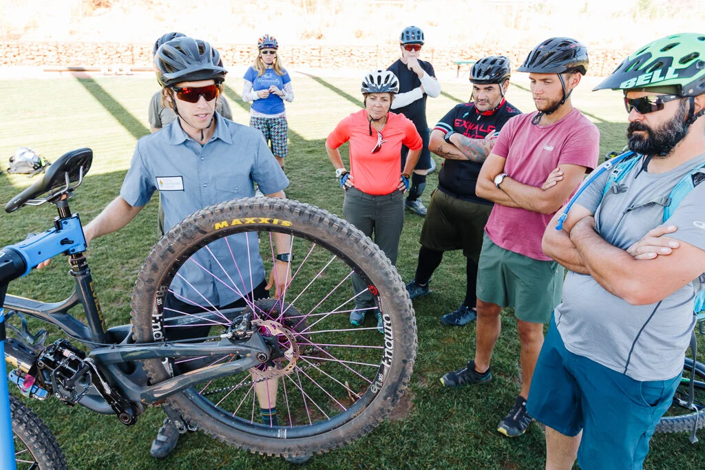 Group of mountain biking enthusiasts, wearing helmets and casual wear, gathered around a person explaining bike maintenance outdoors on a sunny day.
