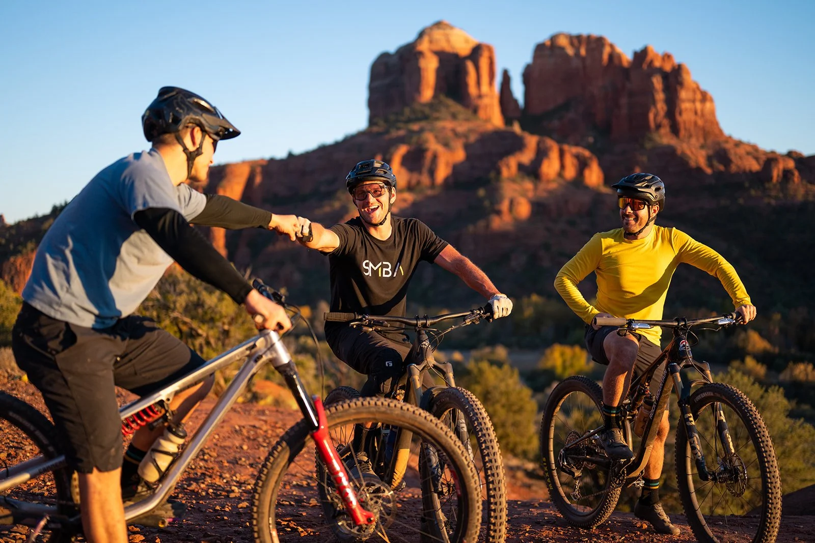 Three men riding mountain bikes in a desert landscape with red rock formations in the background, exchanging a fist bump and smiling.
