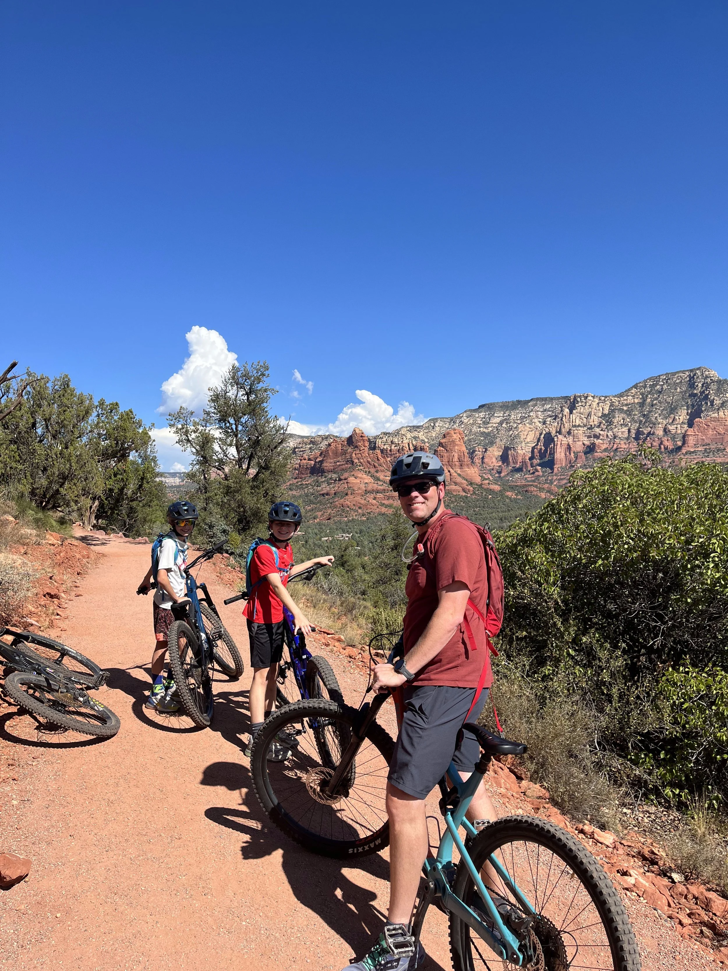 Three people wearing helmets with mountain bikes on a dirt trail in a desert landscape with red rock formations and blue sky.