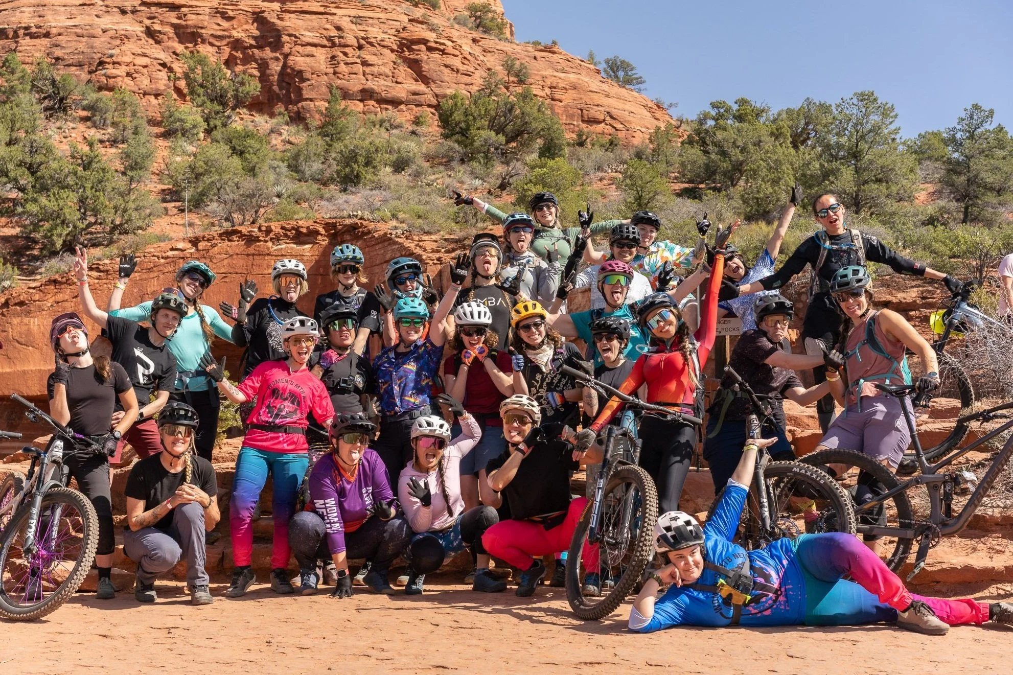 A large group of women wearing cycling gear and helmets, posing with their bikes in front of red rock formations and desert vegetation under a clear blue sky. They are smiling, making peace signs, and celebrating their outdoor biking adventure.